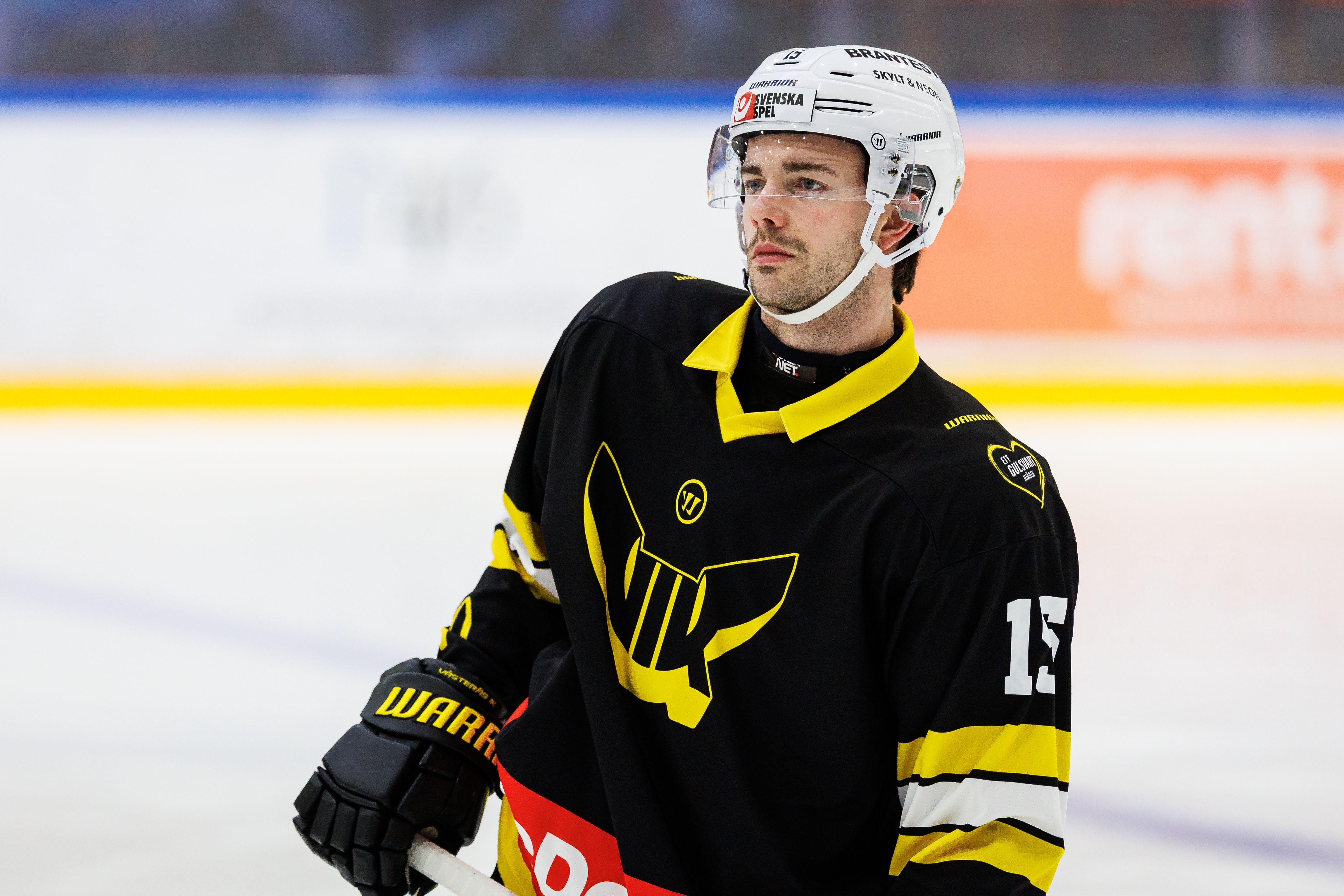 Ishockeyspelare i svart och gul tröja med nummer 13, utrustad med vit hjälm och handskar, står på isen under en match.
