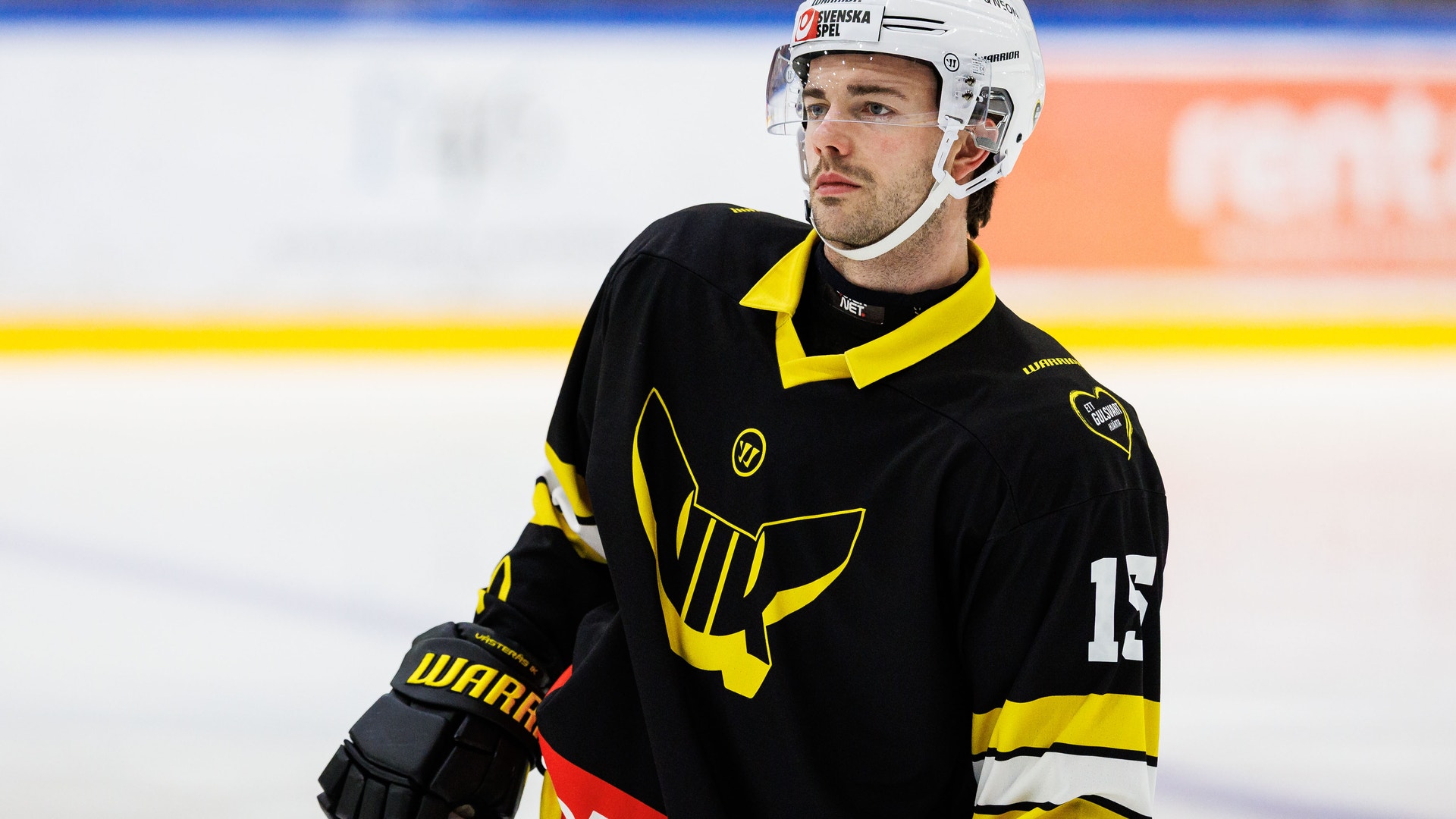 Ishockeyspelare i svart och gul tröja med nummer 13, utrustad med vit hjälm och handskar, står på isen under en match.