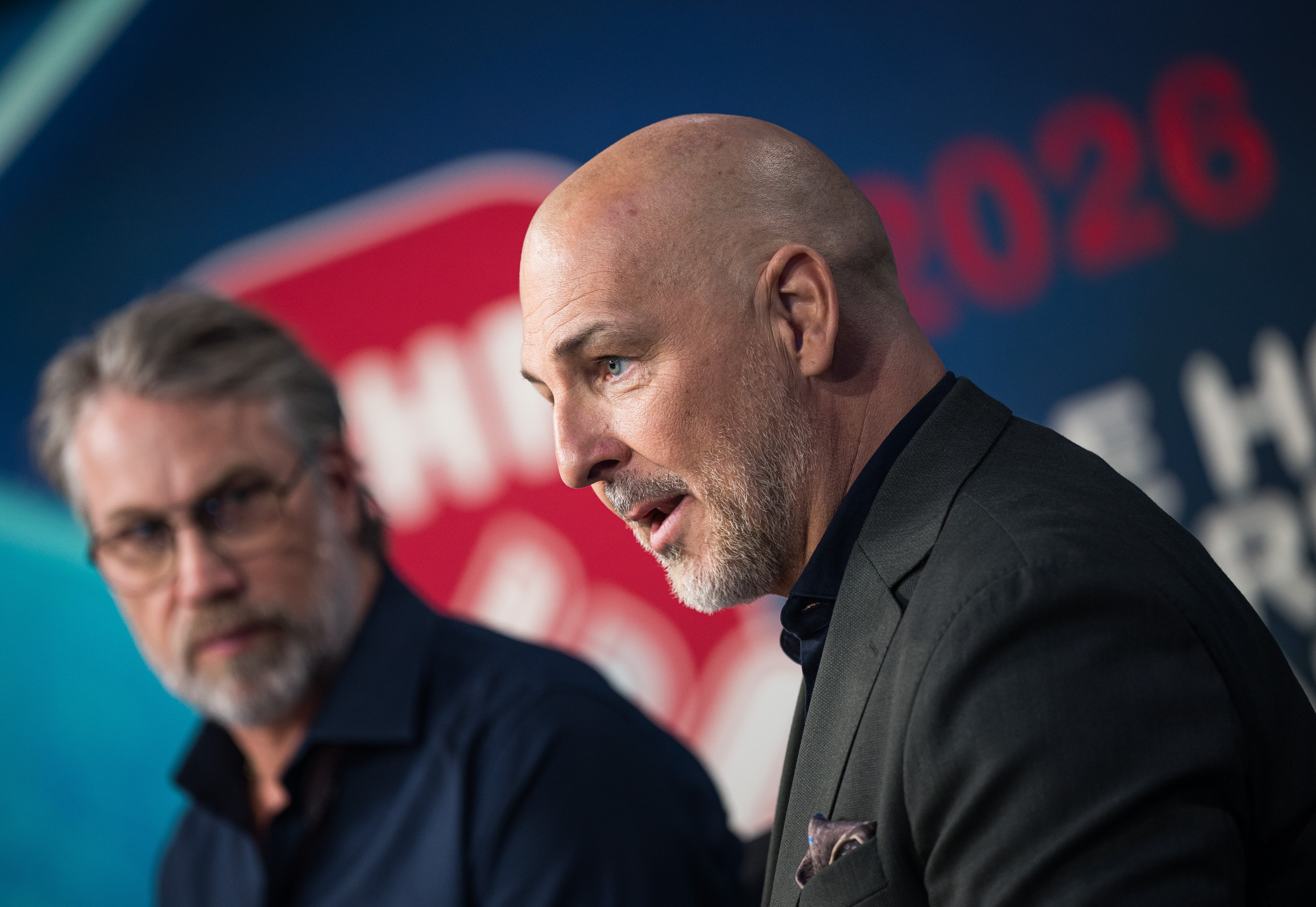 Två män i en intervjusituation framför en vägg med blå och röd belysning. En man i svart blazer talar och gestikulerar medan den andra lyssnar.

Ishockeyspelare i svart och vit tröja gestikulerar under en match. Spelaren bär hjälm och är fotograferad från sidan med publik i suddig bakgrund.