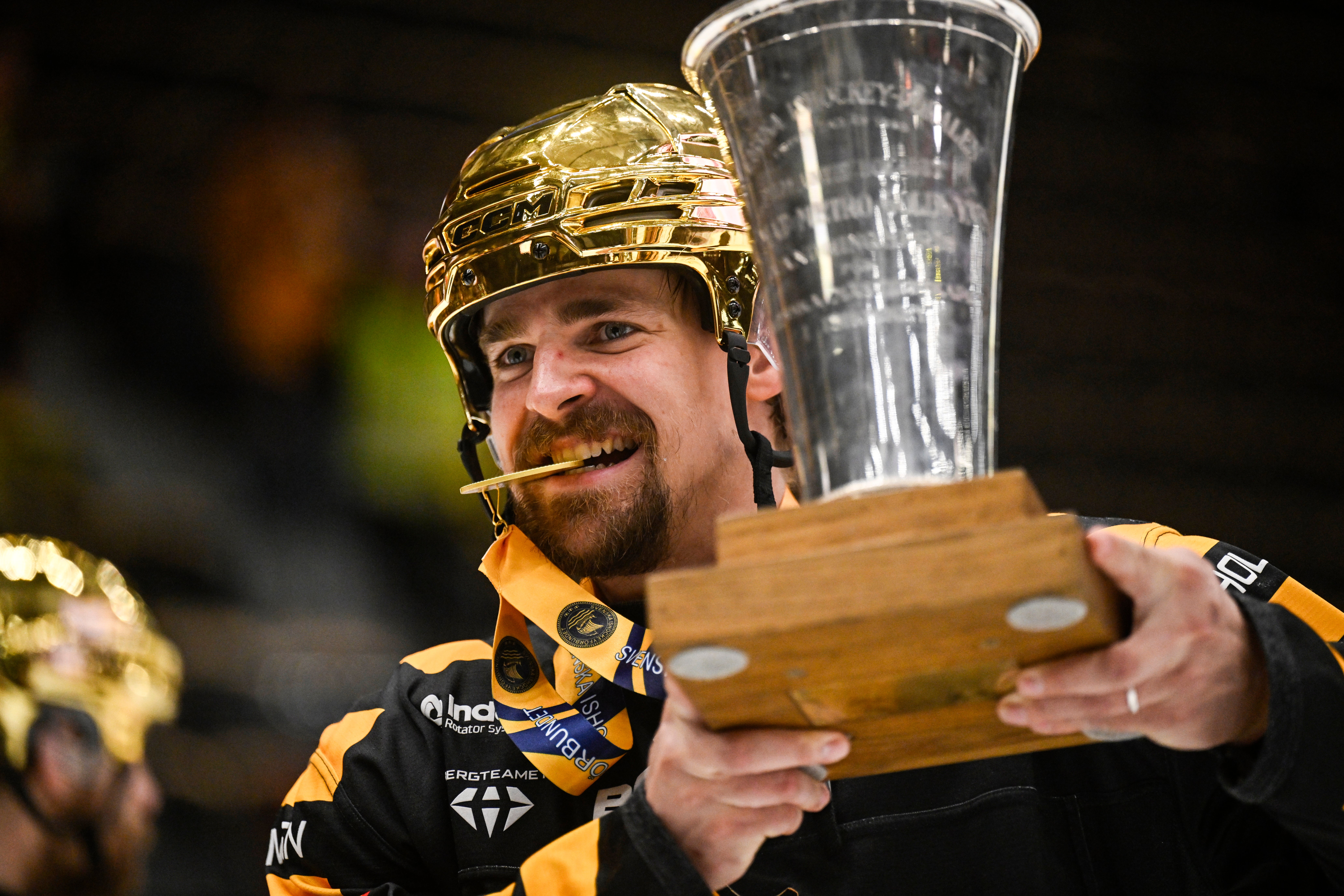 En glädjestrålande hockeyspelare i gul och svart utrustning håller en pokal och ett hockeykryp efter en seger.