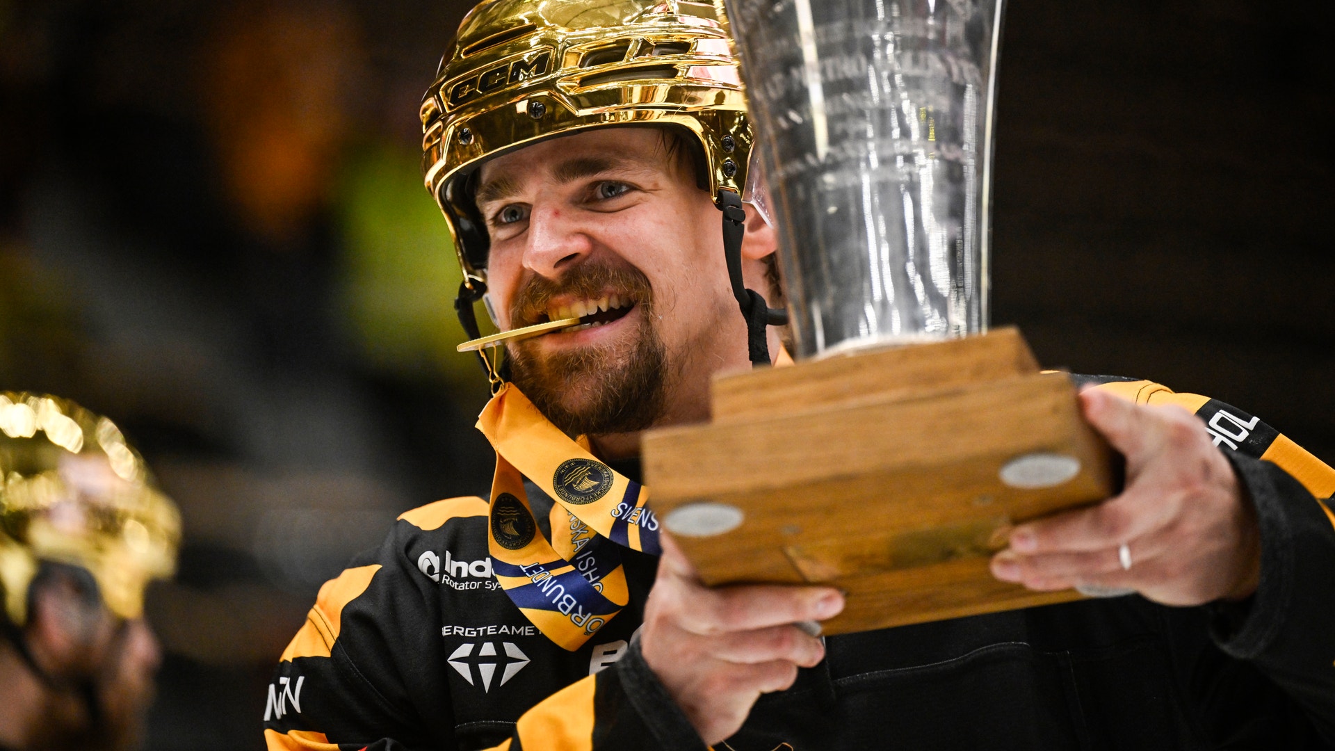 En glädjestrålande hockeyspelare i gul och svart utrustning håller en pokal och ett hockeykryp efter en seger.