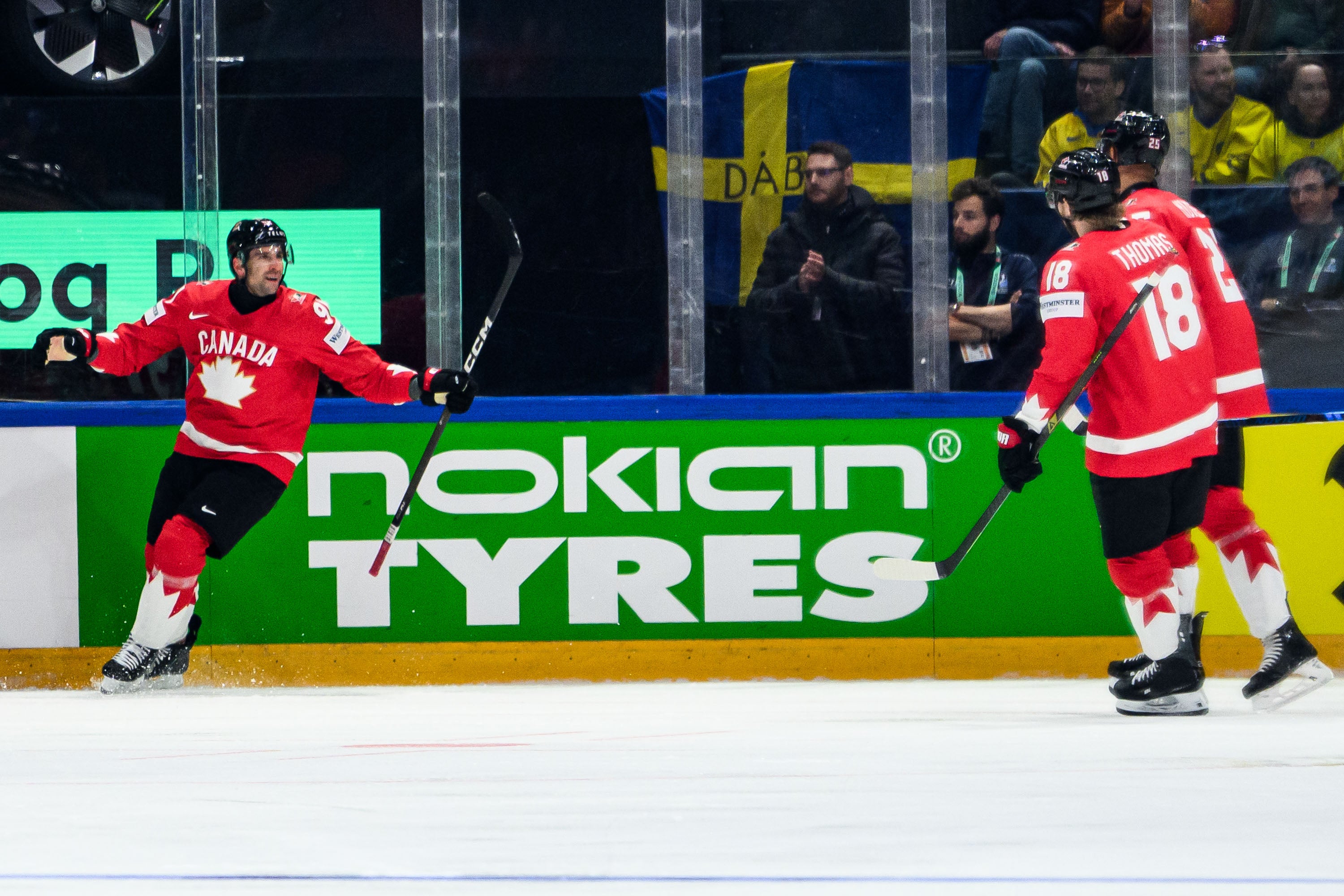 Två ishockeyspelare i röda kanadensiska landslagströjor spelar på isen under en match. I bakgrunden syns sponsorlogotyper och åskådare.