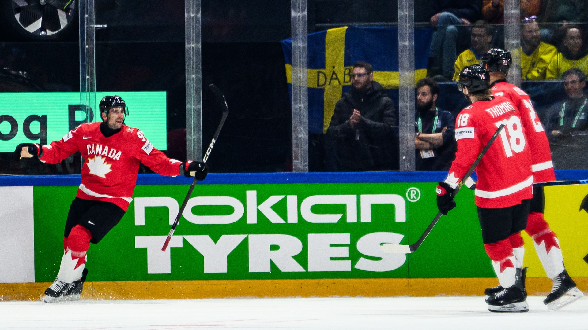 Två ishockeyspelare i röda kanadensiska landslagströjor spelar på isen under en match. I bakgrunden syns sponsorlogotyper och åskådare.