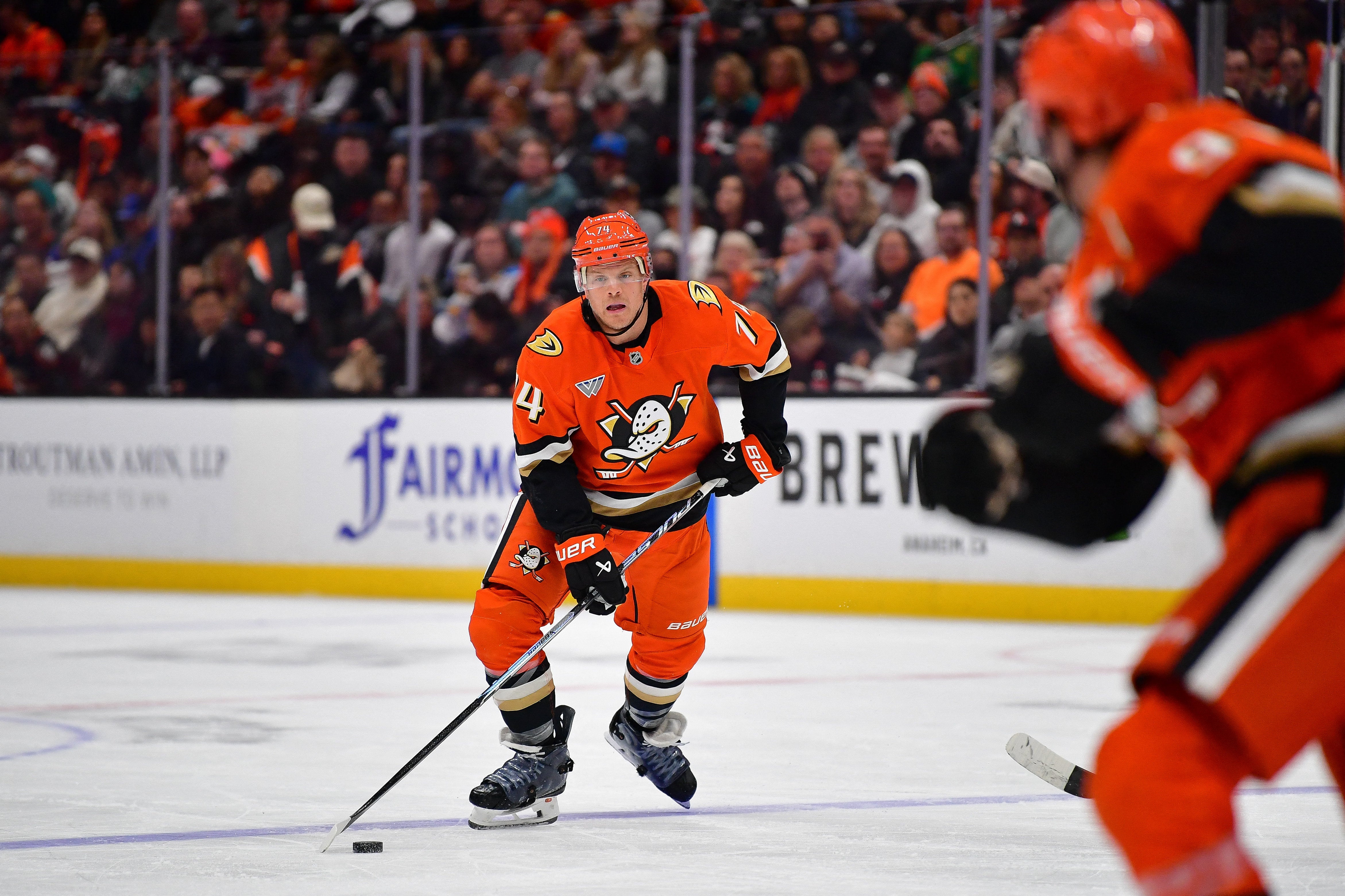 Ishockeyspelare i orange uniform med puck på isen under en match med publik i bakgrunden.