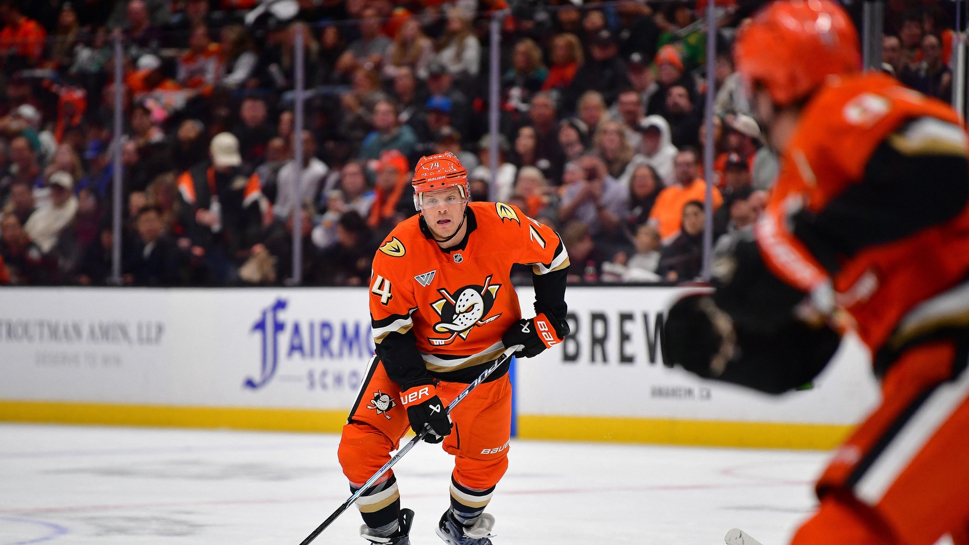 Ishockeyspelare i orange uniform med puck på isen under en match med publik i bakgrunden.