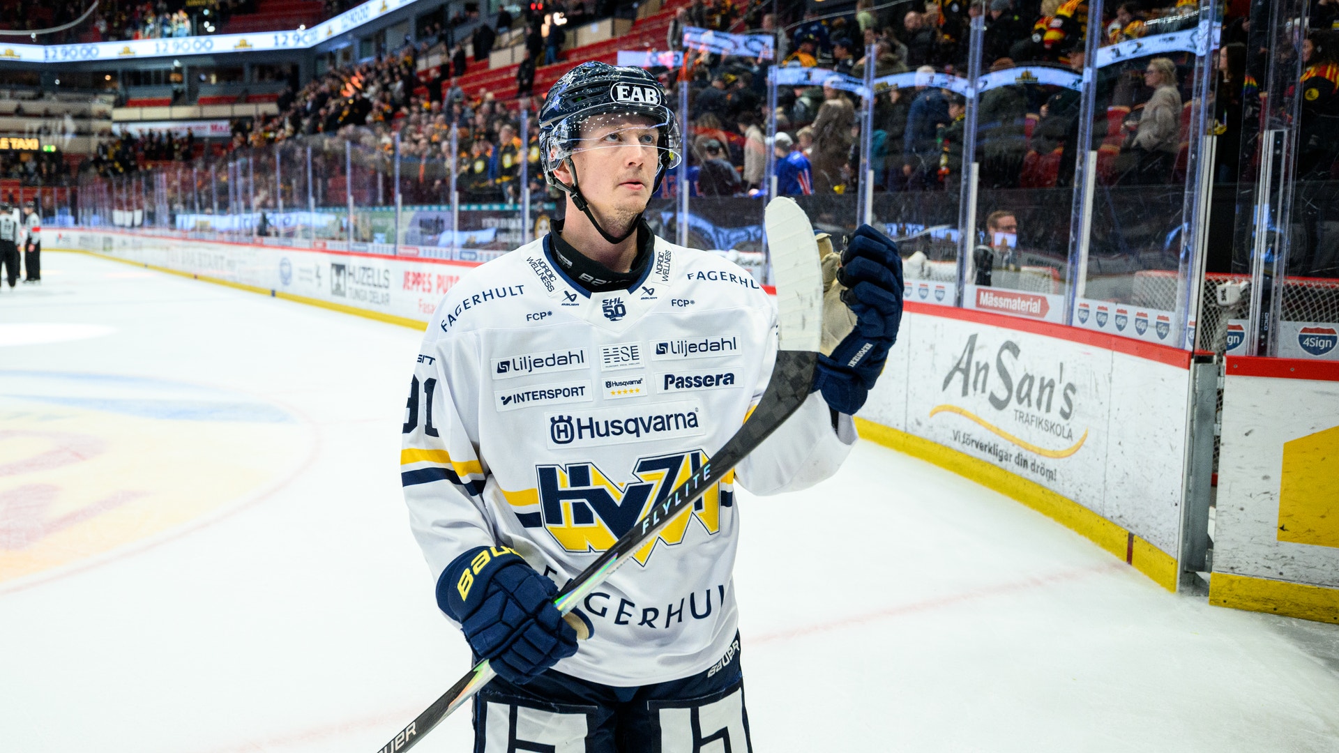 Ishockeyspelare i vit matchdräkt med blå och gula detaljer står på isen under en match i en fullsatt ishall.