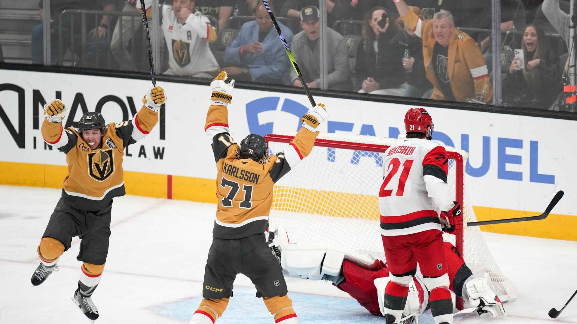 Två ishockeyspelare i gula och svarta Vegas Golden Knights-tröjor jublar med armarna i luften efter ett gjort mål. En motspelare i röd och vit tröja står vid målet. Publiken syns i bakgrunden bakom glaset.