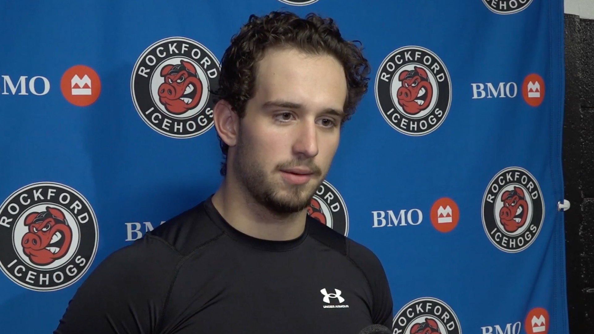 Porträttbild av en man i svart Under Armour-tröja framför en blå sponsorbakgrund med Rockford IceHogs och BMO-logotyper.
