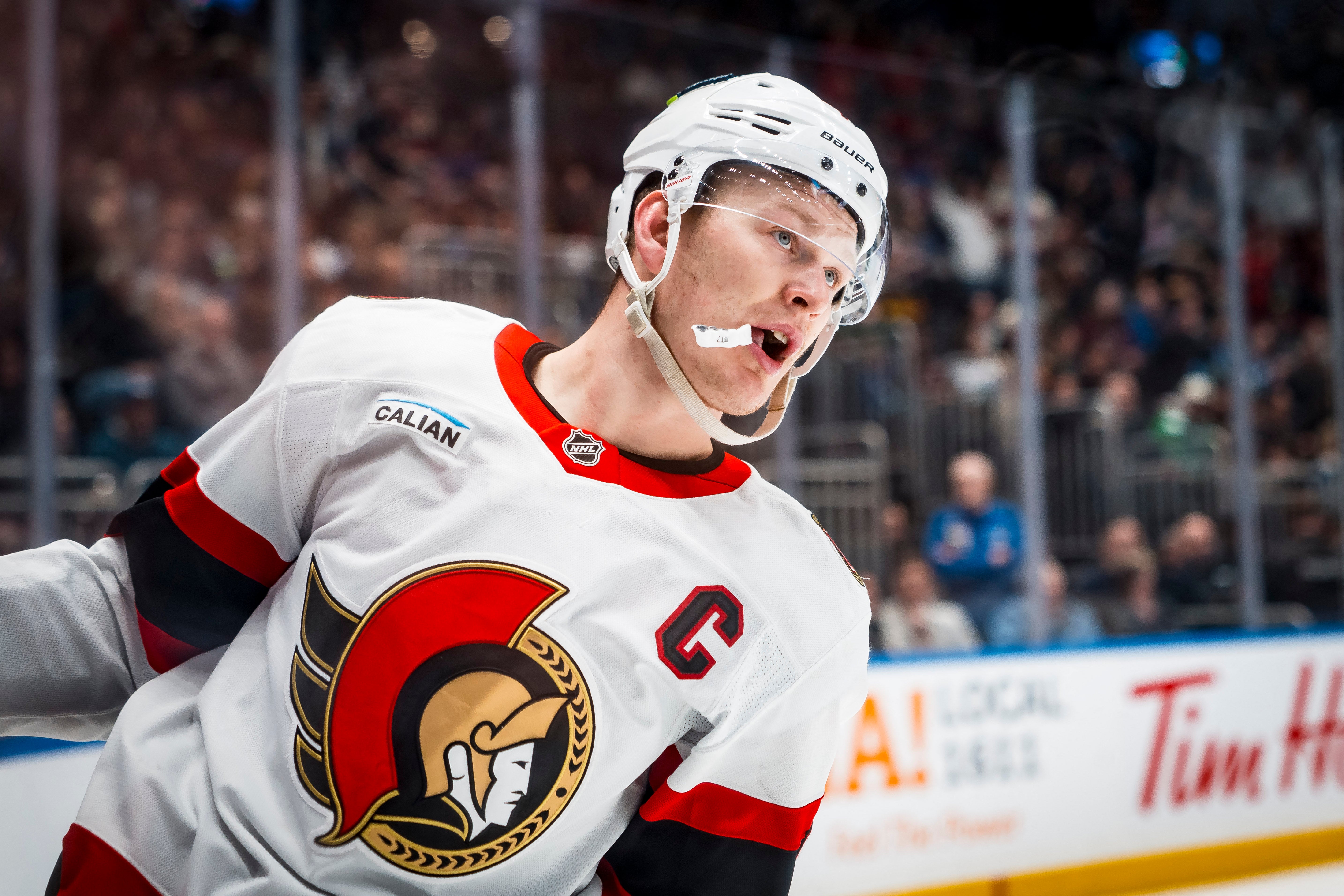 En ishockeyspelare i Ottawa Senators vita tröja under en match. Spelaren bär vit hjälm och ser ut att vara fokuserad på spelet. Publiken syns suddig i bakgrunden.