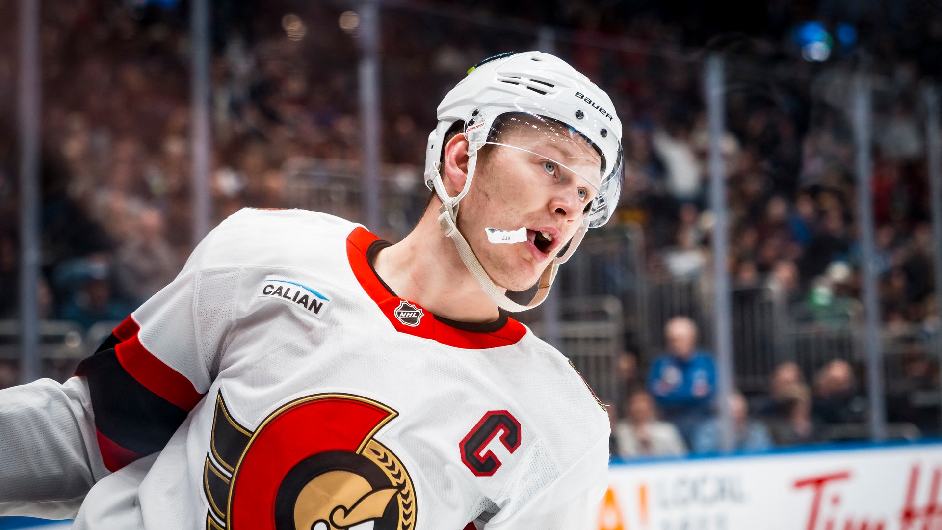 En ishockeyspelare i Ottawa Senators vita tröja under en match. Spelaren bär vit hjälm och ser ut att vara fokuserad på spelet. Publiken syns suddig i bakgrunden.