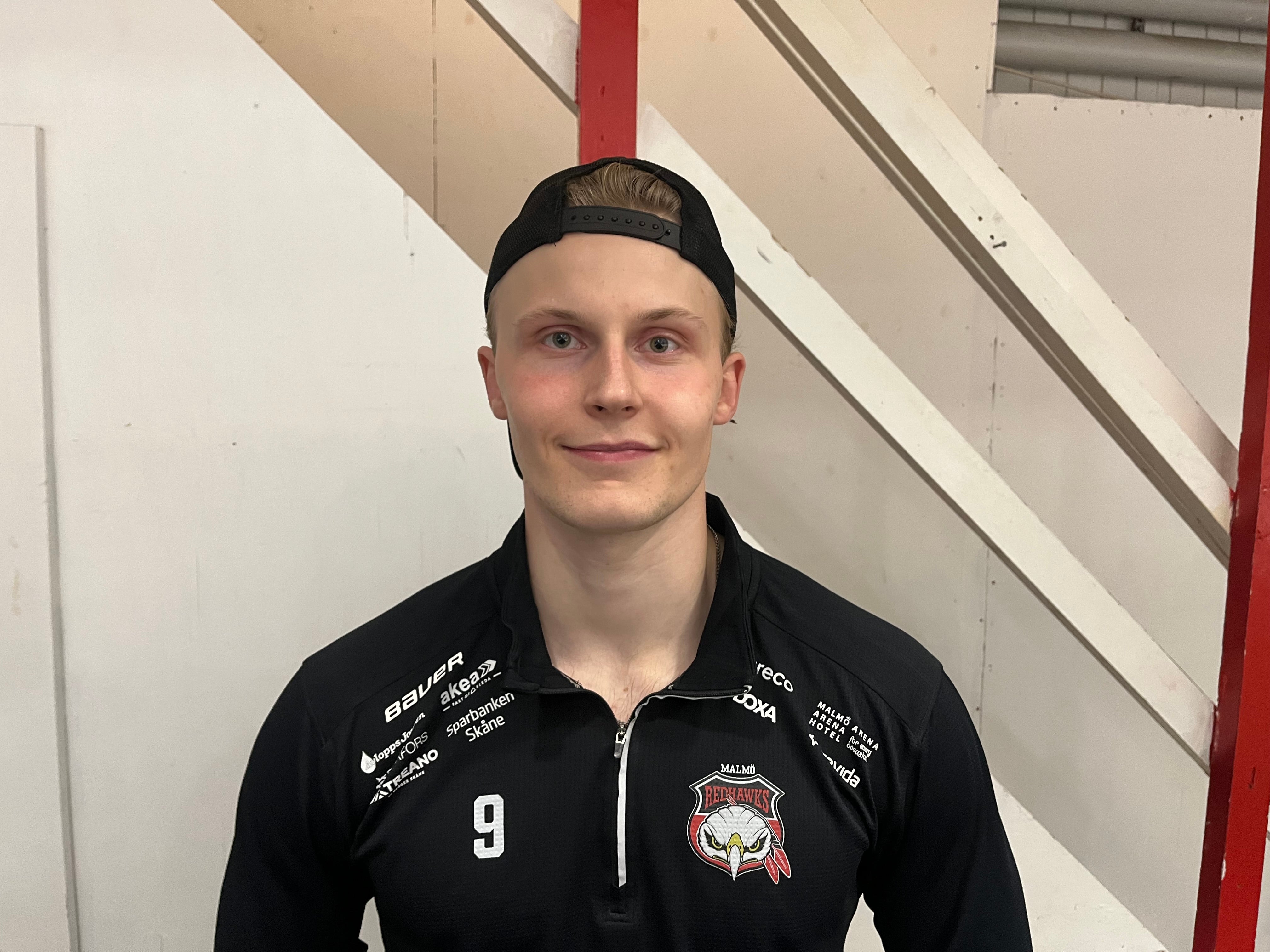 En ung man i svart tröja och keps ler mot kameran. En ung ishockeyspelare i svart tröja med Malmö-emblem och matchtröjenummer 9 ler in i kameran, med en keps baklänges på huvudet.