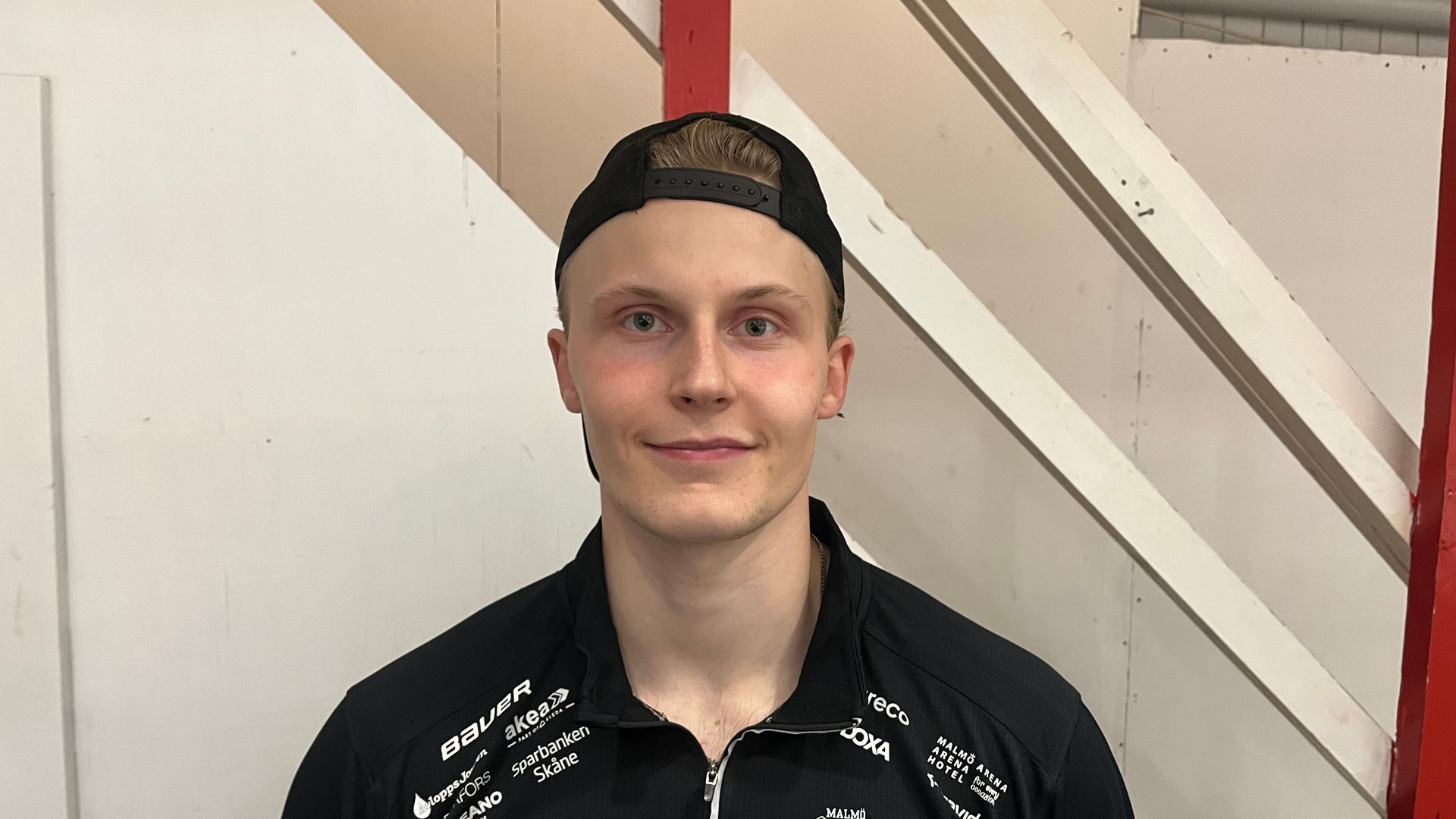 En ung man i svart tröja och keps ler mot kameran. En ung ishockeyspelare i svart tröja med Malmö-emblem och matchtröjenummer 9 ler in i kameran, med en keps baklänges på huvudet.