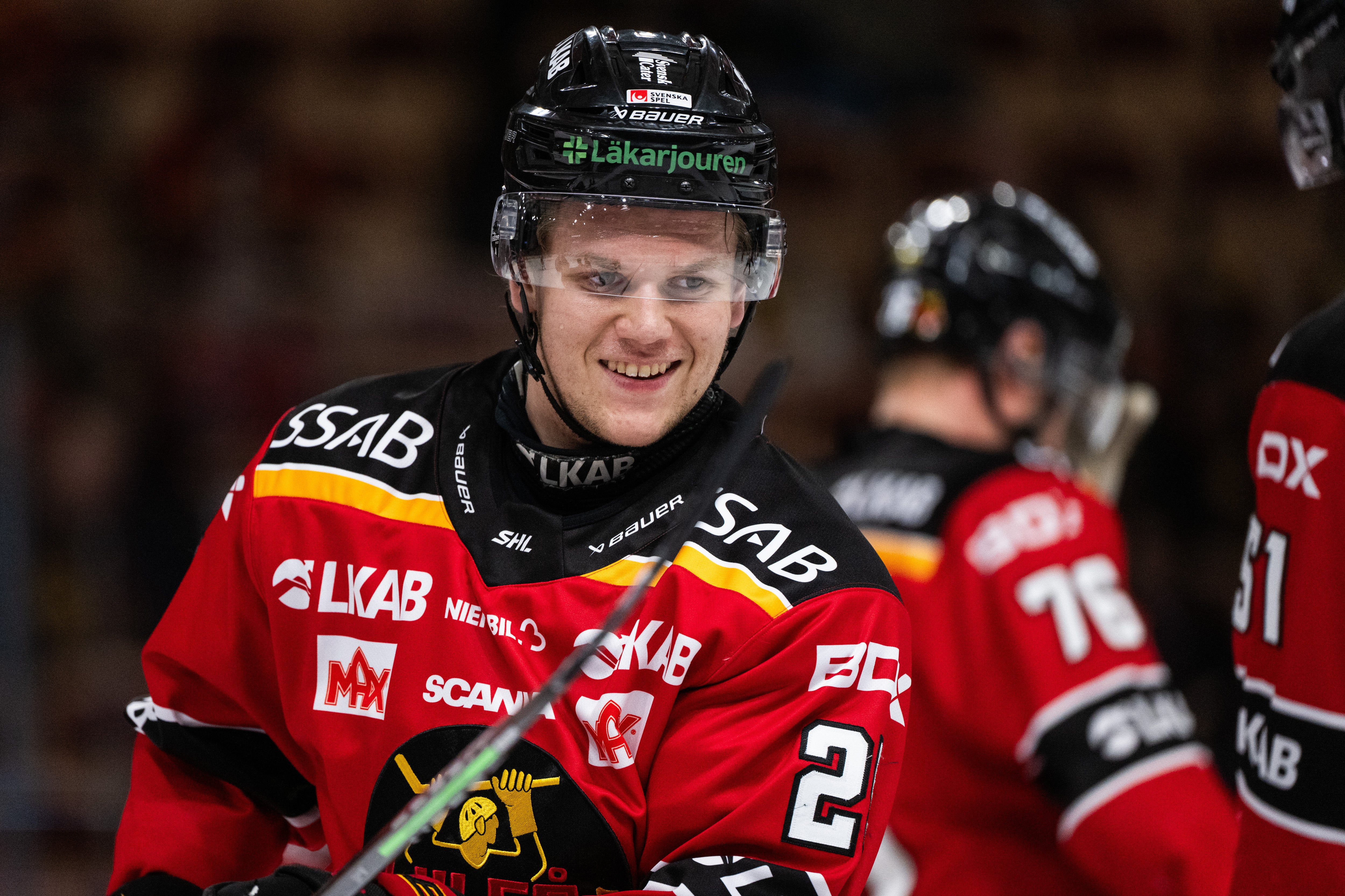 Ishockeyspelare i röd och svart matchdräkt ler mot kameran under en ishockeymatch.