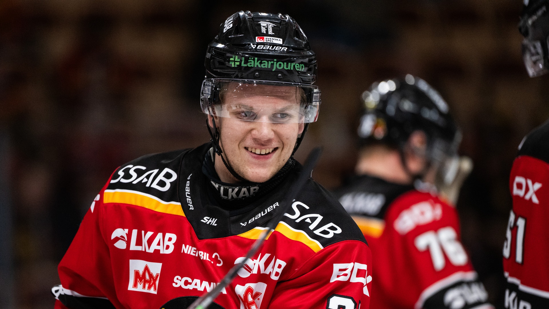 Ishockeyspelare i röd och svart matchdräkt ler mot kameran under en ishockeymatch.