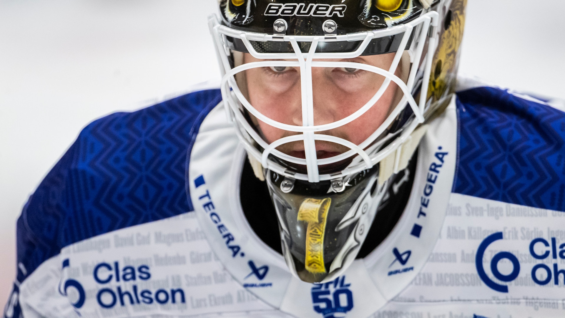 Porträtt av en ishockeysmålvakt i blå och vit uniform med skyddsutrustning och ansiktsmask.