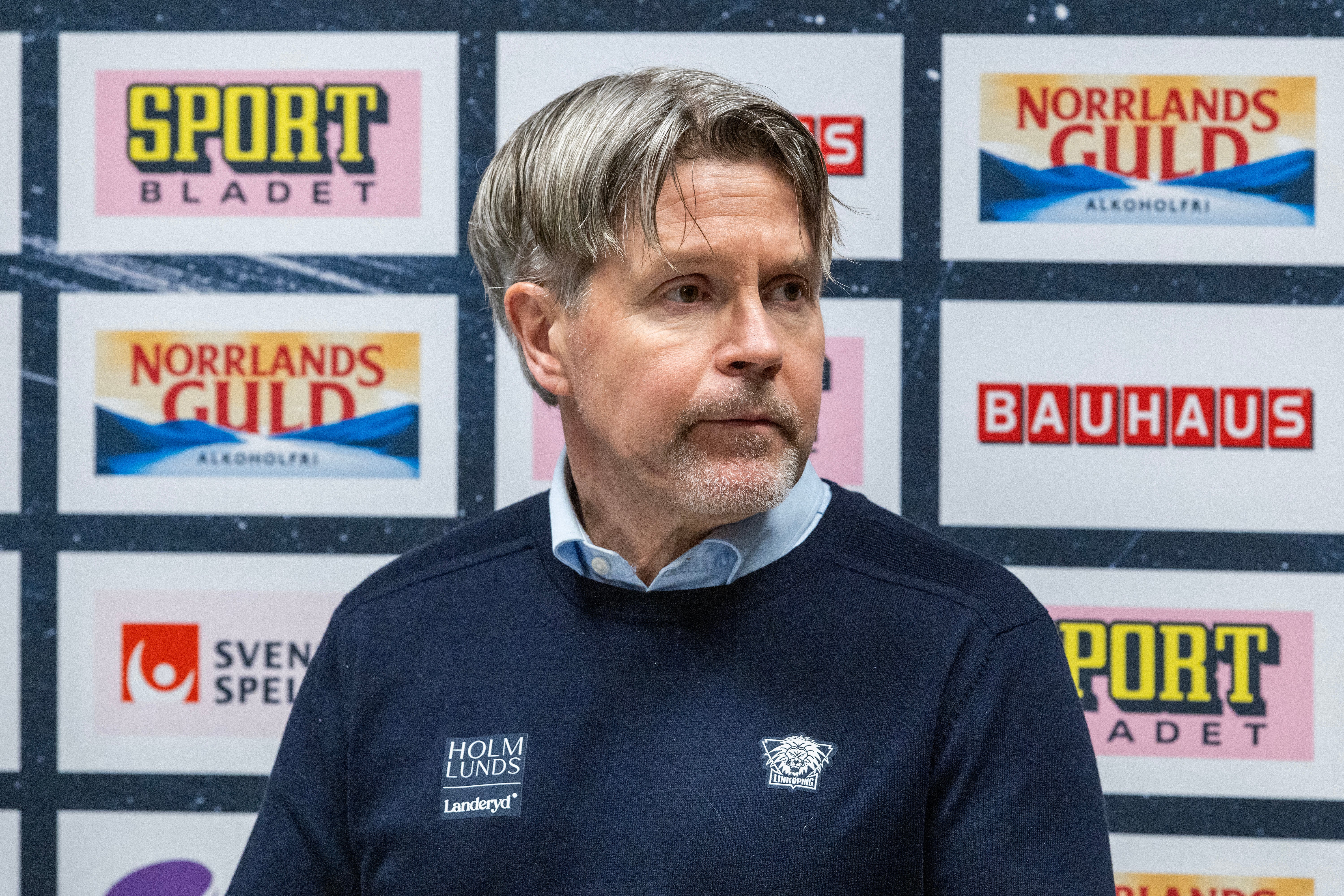 En man i mörk tröja framför en pressvägg med sponsorlogotyper.