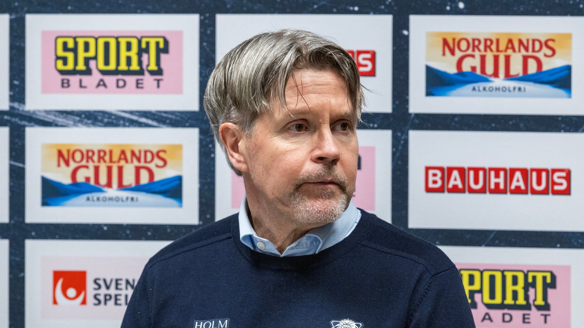 En man i mörk tröja framför en pressvägg med sponsorlogotyper.
