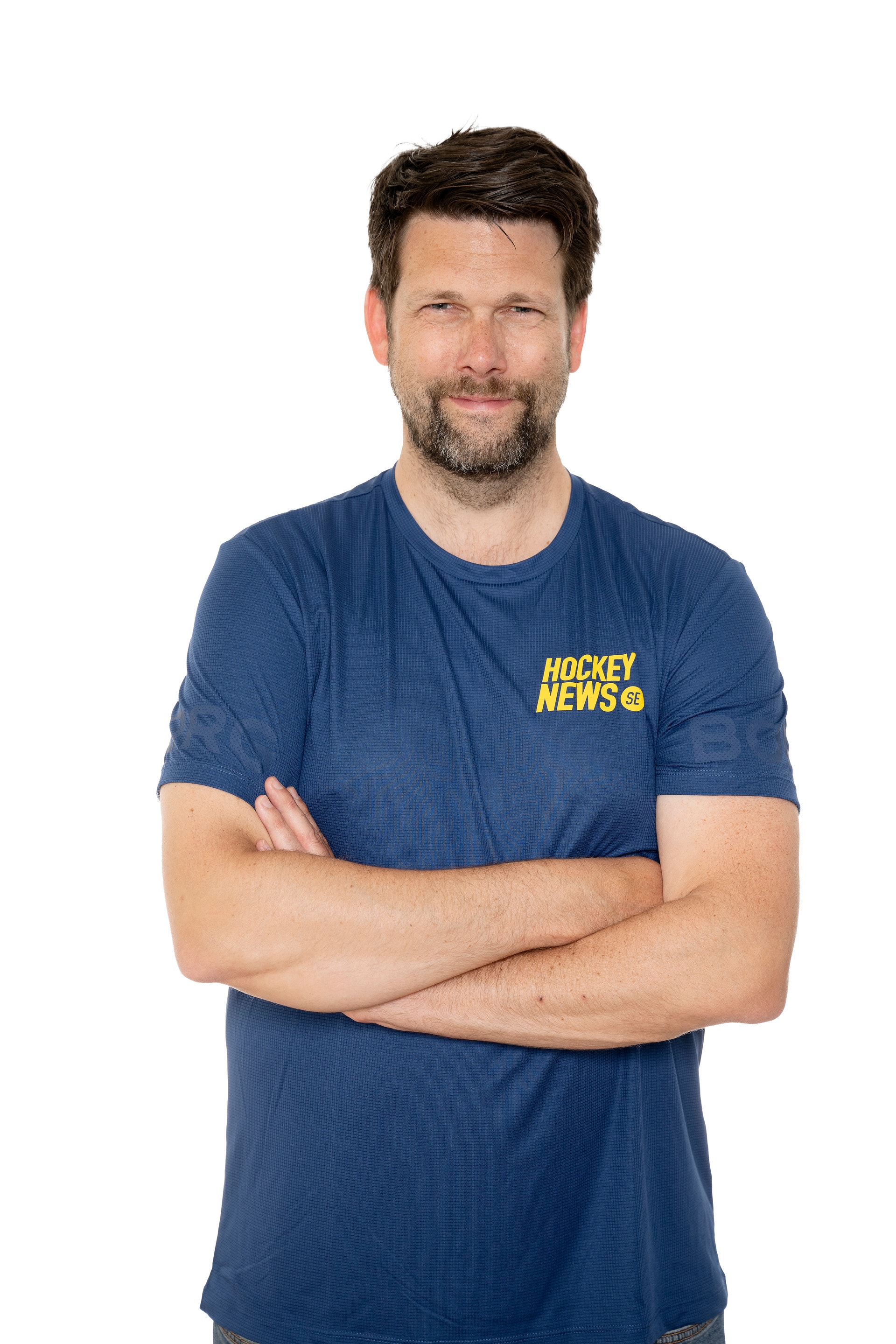 Porträtt av en man i mörkblå t-shirt med Hockey News logga, armarna korsade, framför vit bakgrund.