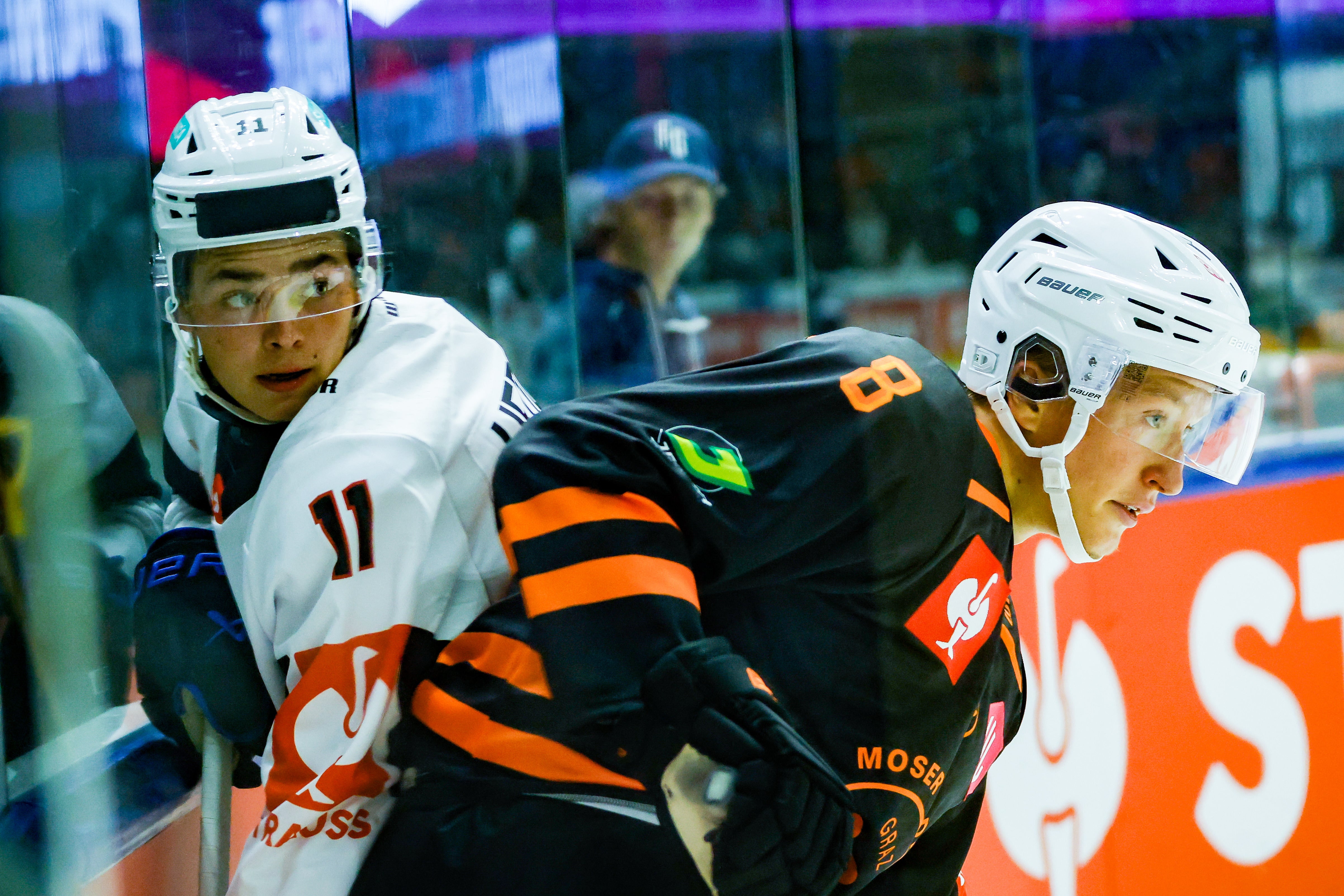 Två ishockeyspelare i hjälmar och matchtröjor, en i vitt och en i svart och orange, står nära avbalkningen under en match. I bakgrunden syns glasskivan och en åskådare.