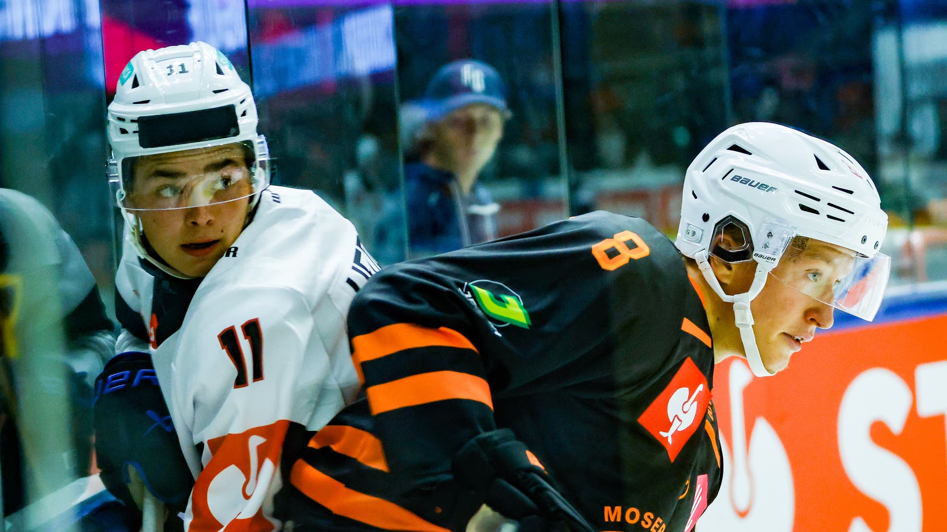 Två ishockeyspelare i hjälmar och matchtröjor, en i vitt och en i svart och orange, står nära avbalkningen under en match. I bakgrunden syns glasskivan och en åskådare.