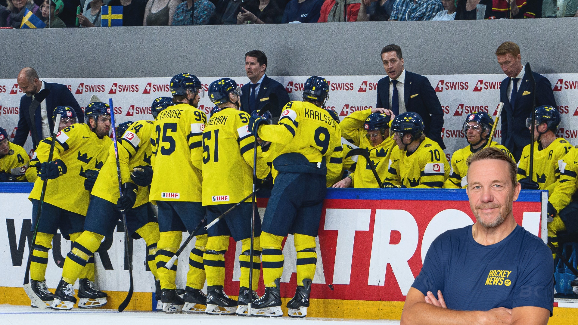 Ishockeylag i gula och svarta trikåer samlat vid bänken under en match. En man i mörkblå t-shirt står i förgrunden med armarna i kors. I bakgrunden syns publik och reklamskyltar.