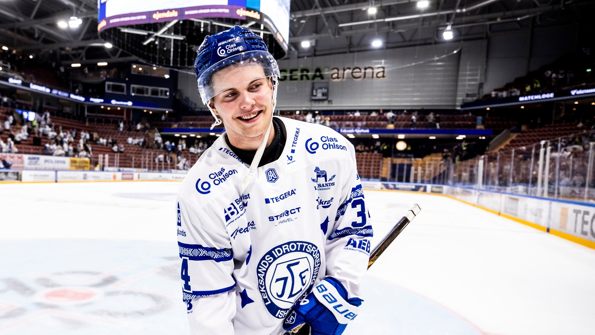 En leende ishockeyspelare i vit matchtröja står på isen i en arena och håller i en klubba.