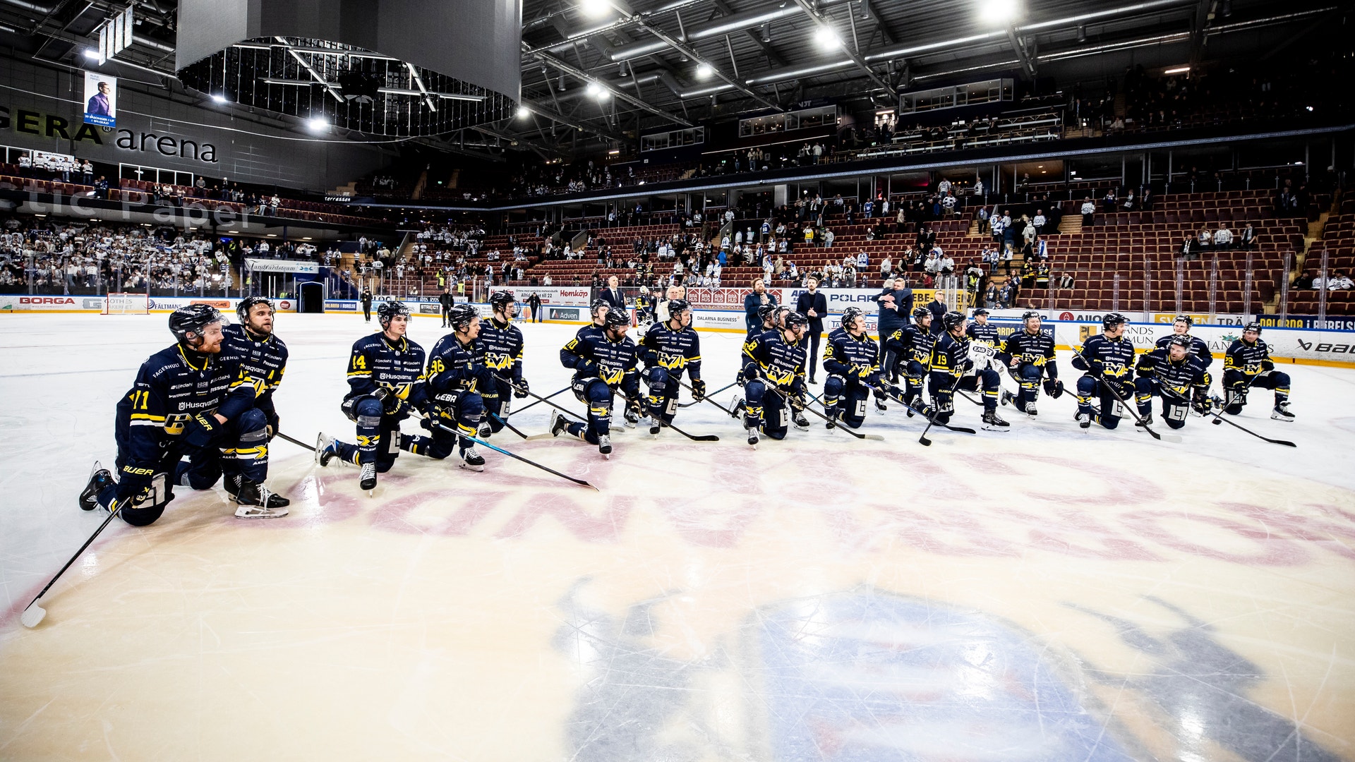 Ishockeymatch i en fullsatt arena. Ett lag i blå och gula trikåer spelar på isen med sina hockeyklubbor. Publiken fyller läktarna i bakgrunden under arenan's ljusa belysning.
