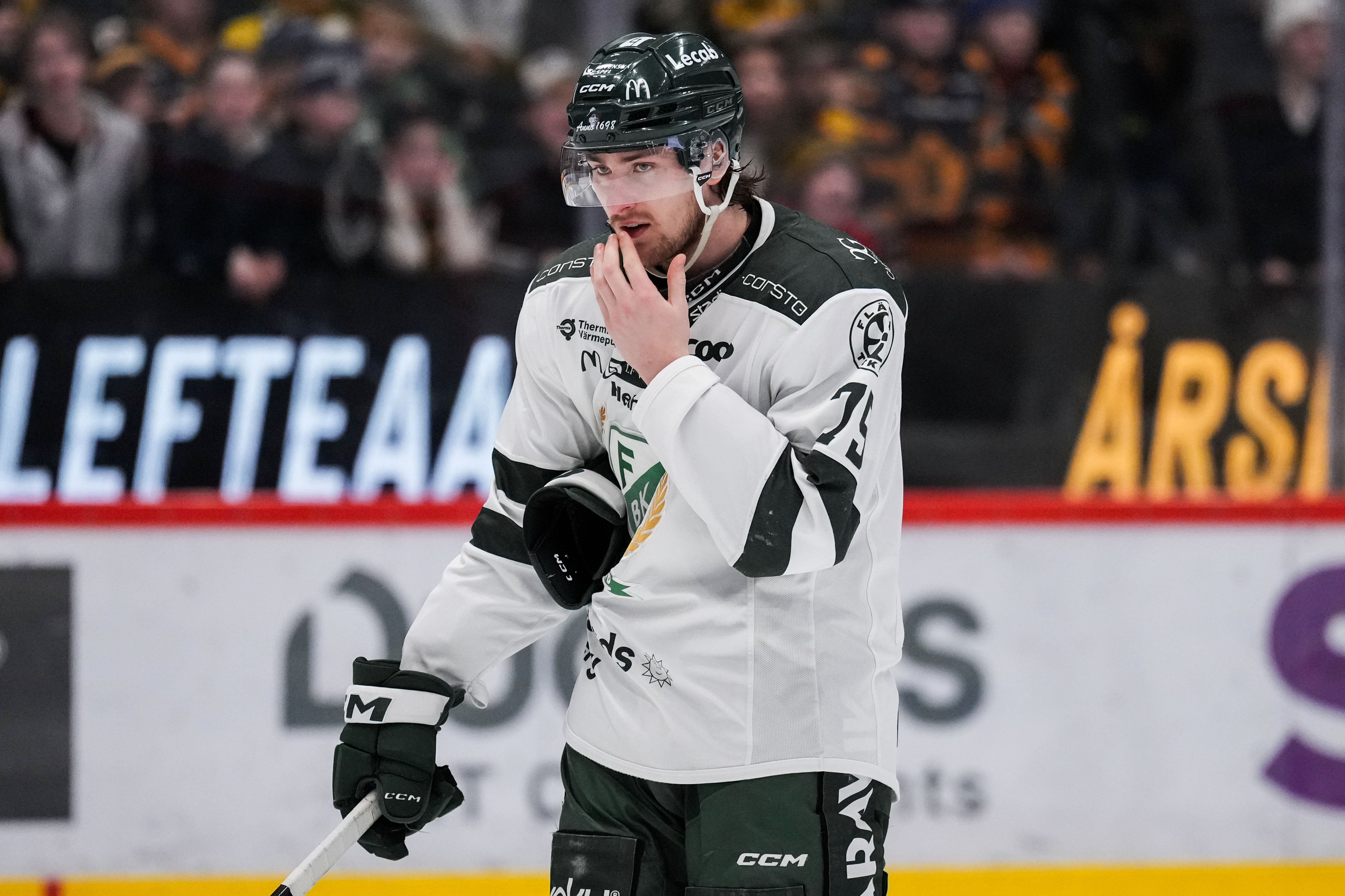 Ishockeyspelare i vit och svart tröja står på isen under en match med handsken mot munnen.