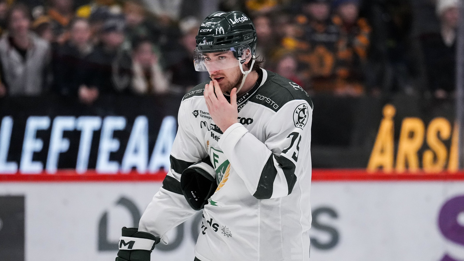 Ishockeyspelare i vit och svart tröja står på isen under en match med handsken mot munnen.