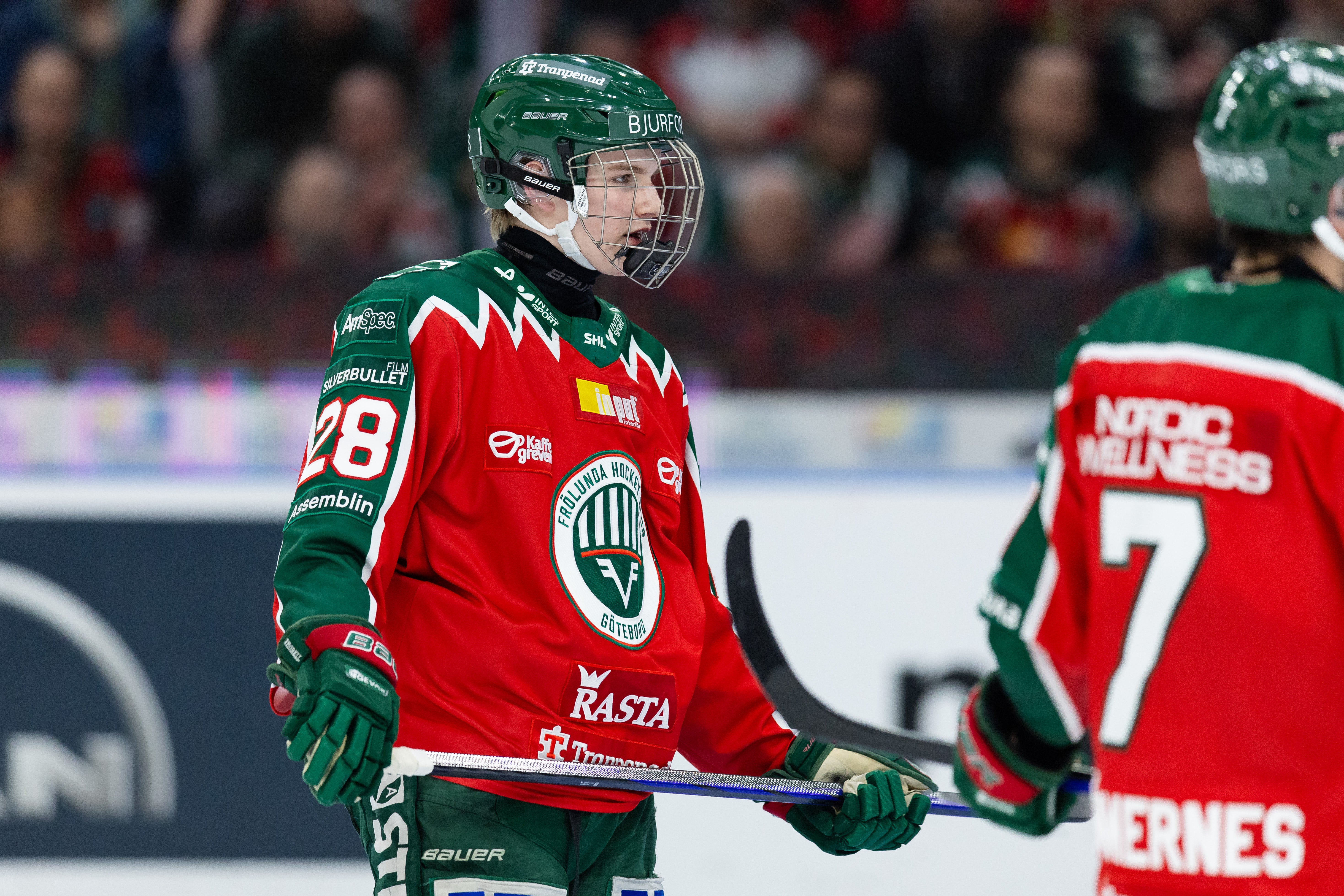 Ishockeyspelare i röd och grön matchdräkt med hjälm och stick under en ishockeymatch.