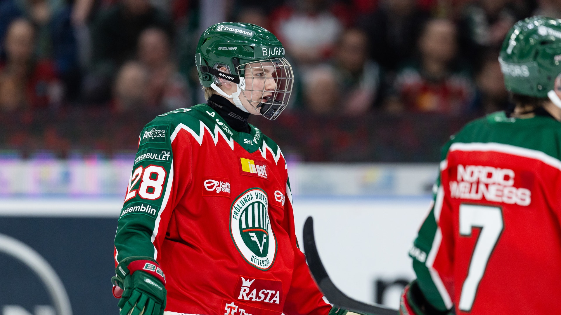 Ishockeyspelare i röd och grön matchdräkt med hjälm och stick under en ishockeymatch.