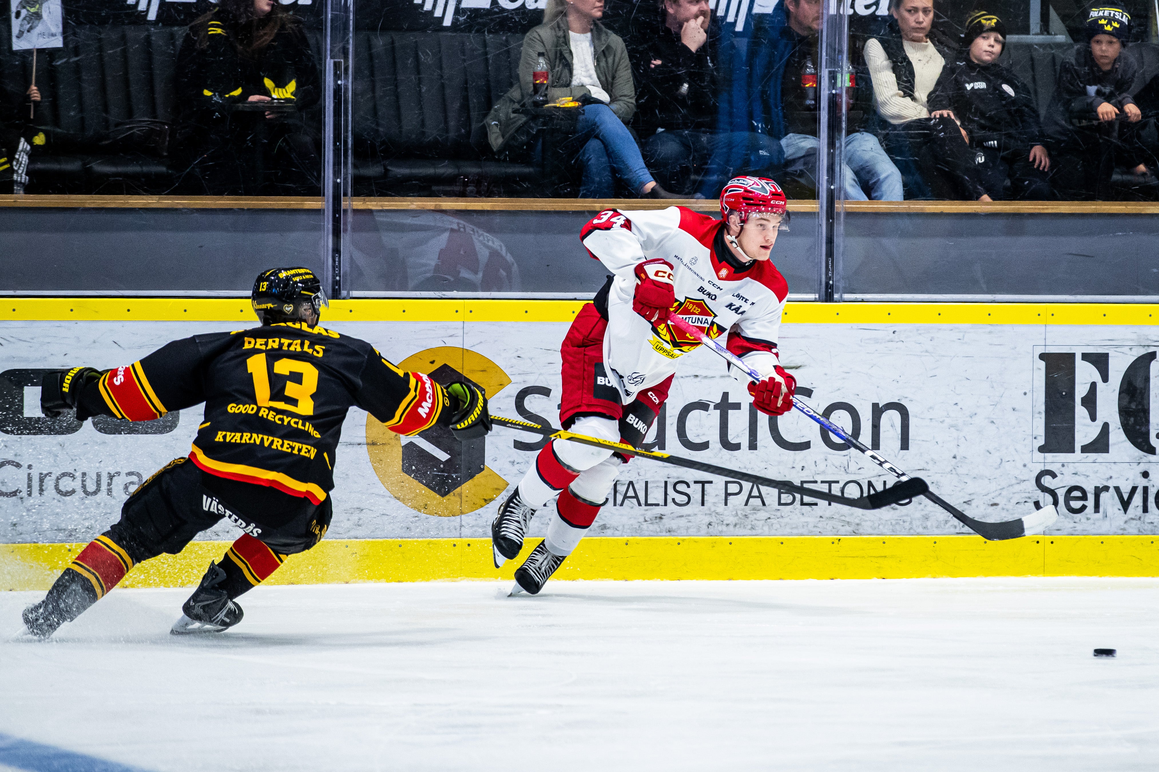 Två ishockeyspelare i matchsituation, en i vit matchtröja och en i svart, med publik i bakgrunden.
