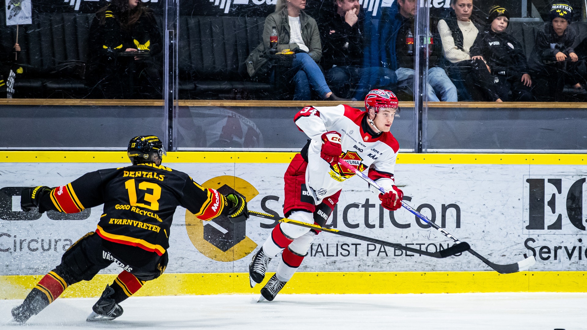 Två ishockeyspelare i matchsituation, en i vit matchtröja och en i svart, med publik i bakgrunden.