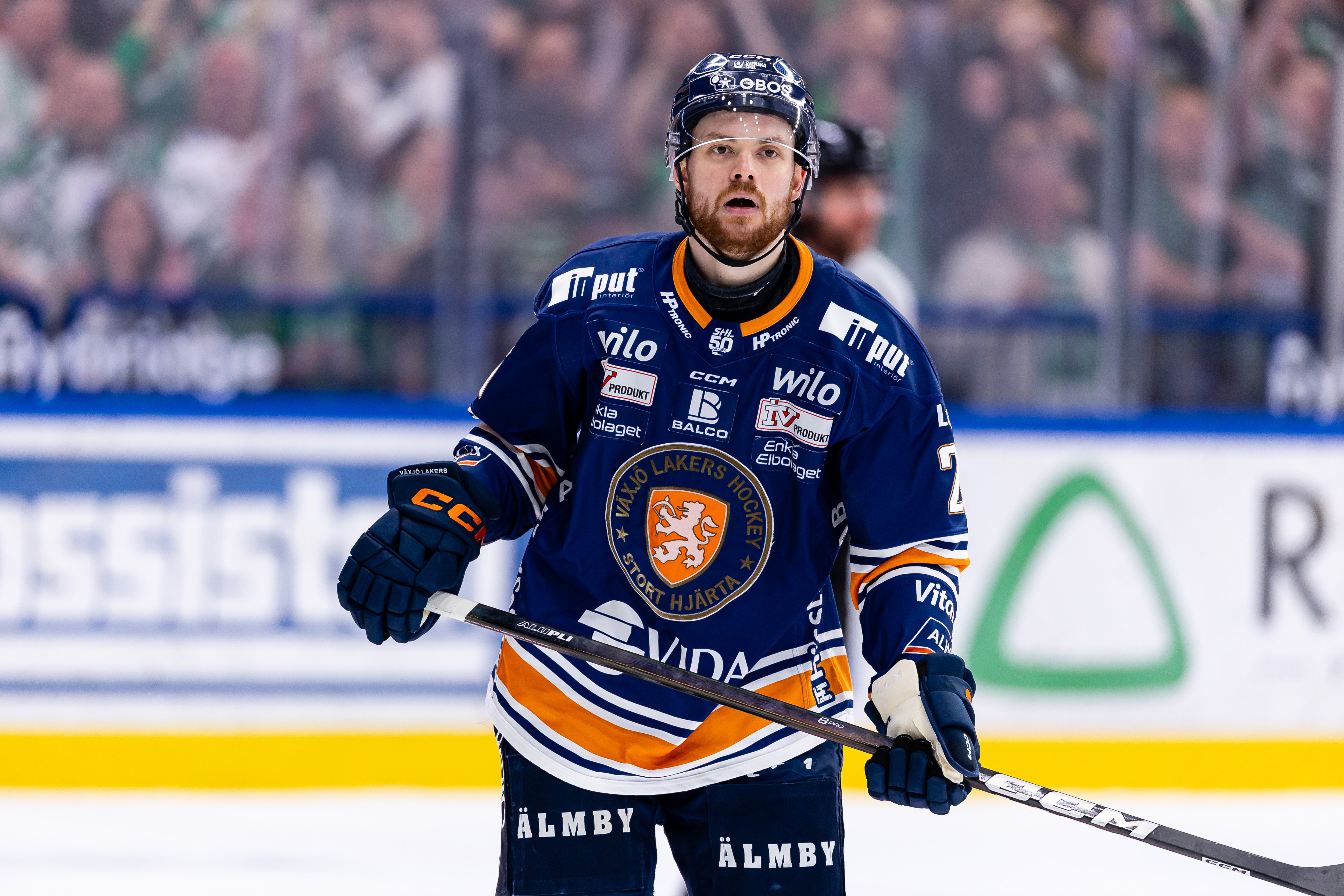 Ishockeyspelare i mörkblå tröja med orange och vita detaljer under en match. Spelaren bär hjälm och handskar och håller en ishockeyklubb. Publiken och reklamskyltar syns i bakgrunden.