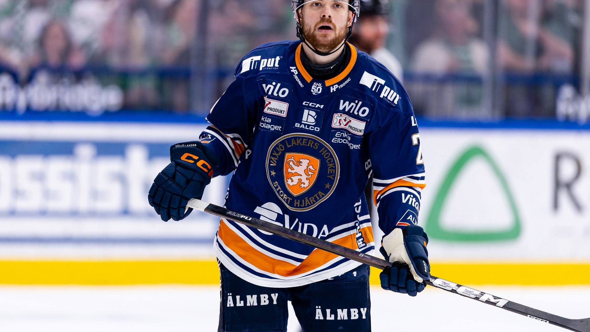 Ishockeyspelare i mörkblå tröja med orange och vita detaljer under en match. Spelaren bär hjälm och handskar och håller en ishockeyklubb. Publiken och reklamskyltar syns i bakgrunden.