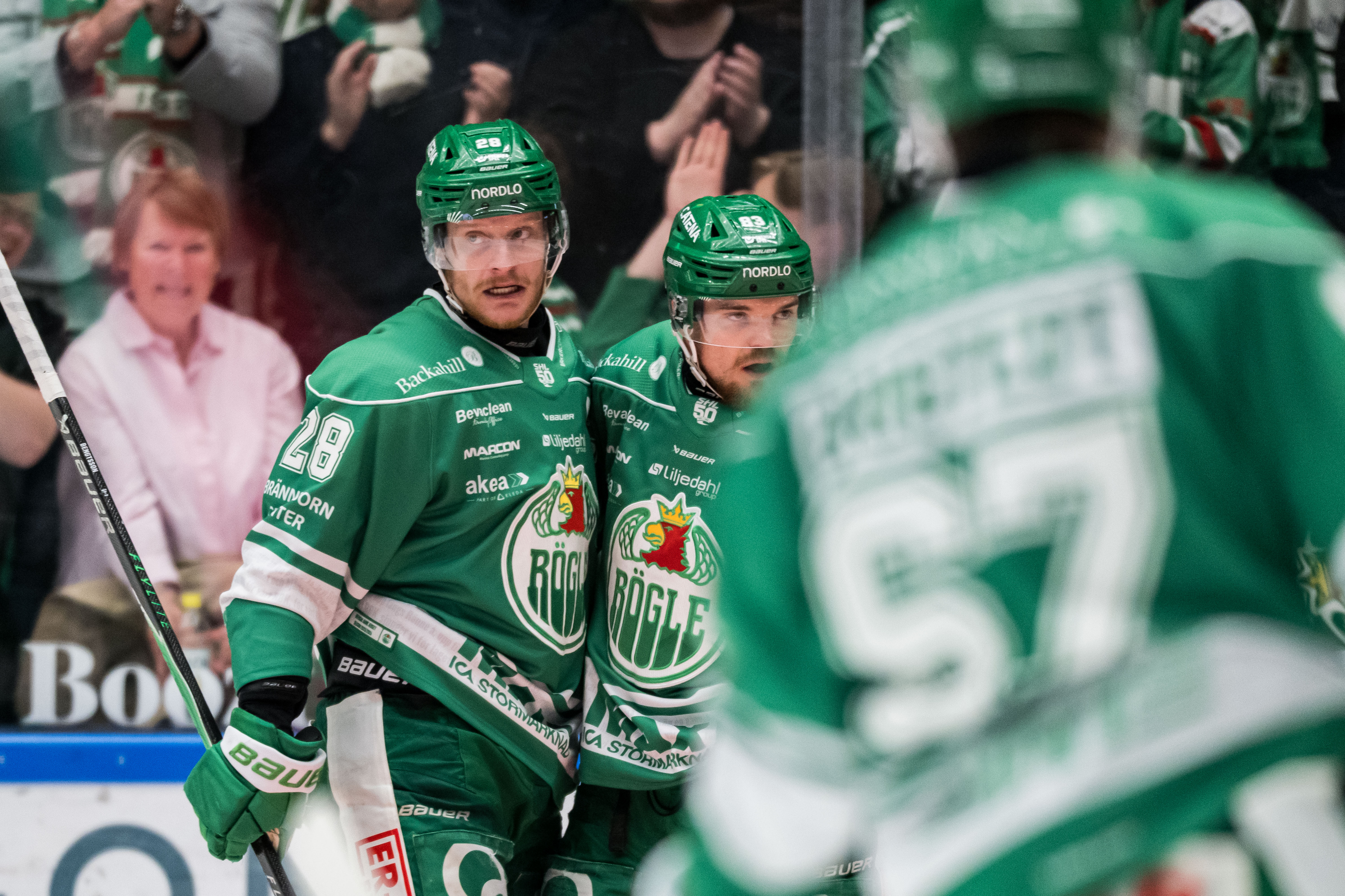 Två ishockeyspelare i gröna tröjor går tillsammans på isen under en match. I bakgrunden syns publik och andra spelare.