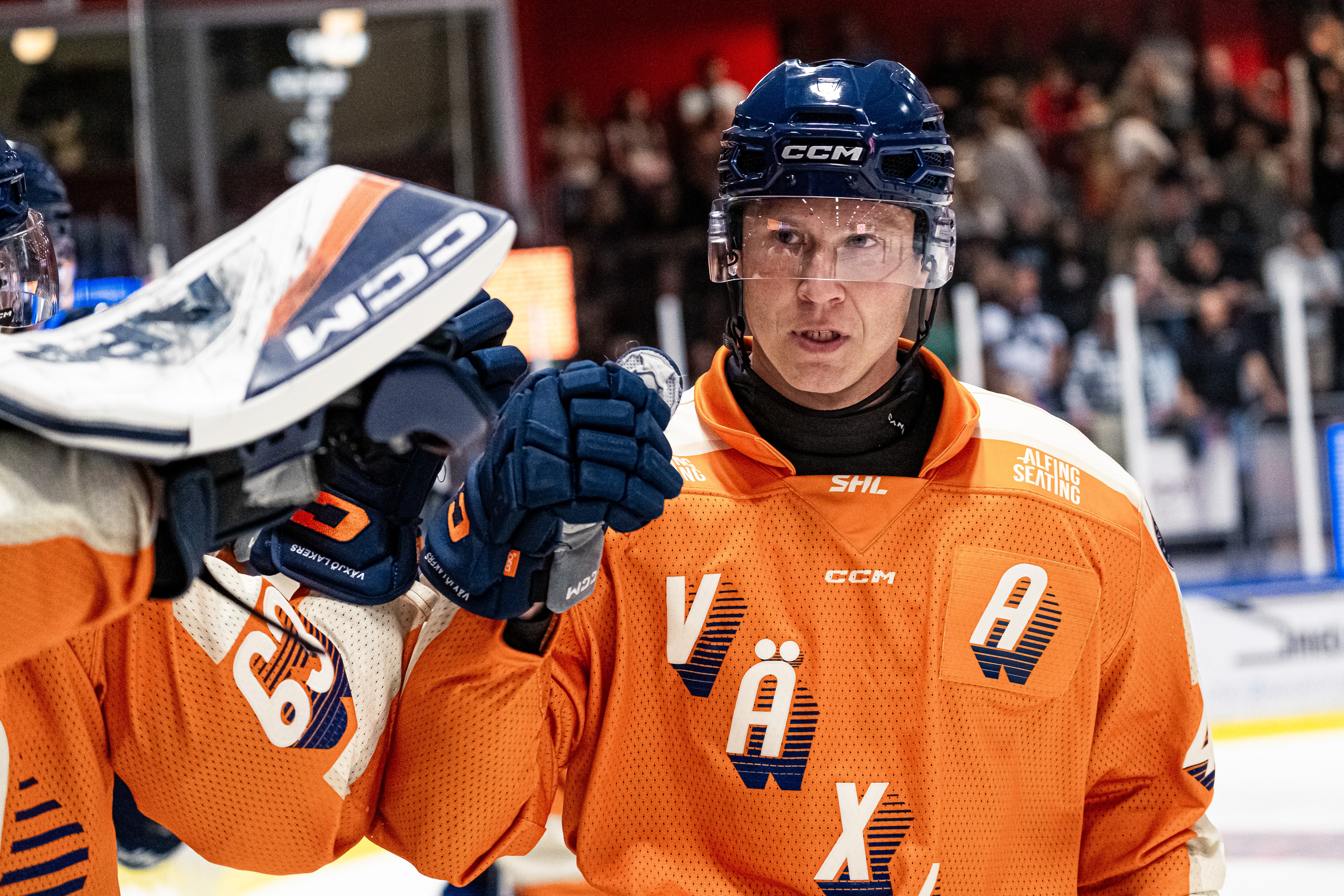 Ishockeyspelare i orange tröja och blå hjälm under en match. Spelaren håller sin ishockeyklubb och tittar mot kameran med publik i bakgrunden.