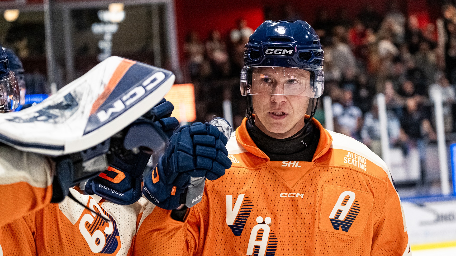 Ishockeyspelare i orange tröja och blå hjälm under en match. Spelaren håller sin ishockeyklubb och tittar mot kameran med publik i bakgrunden.