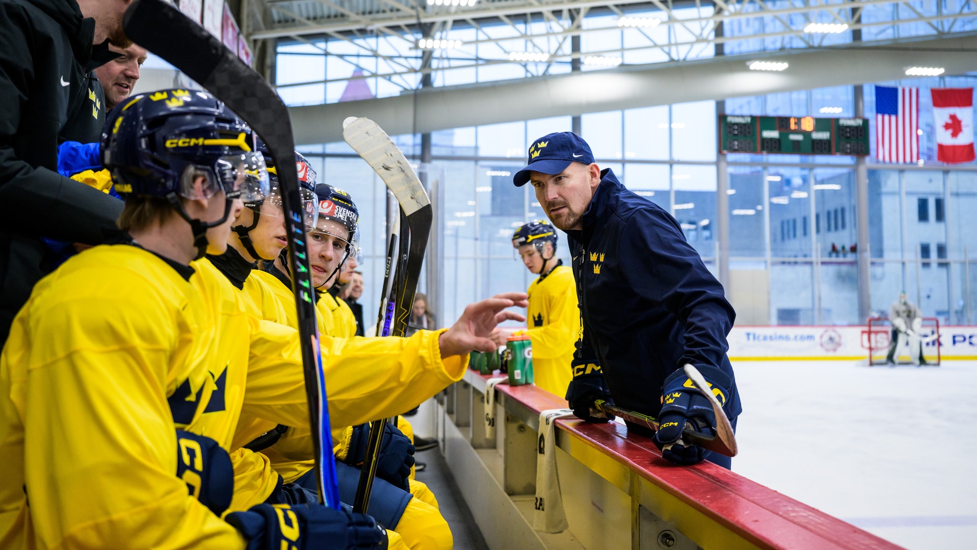 En tränare instruerar en ishockeylag klädda i gula tröjor vid banden på en ishall. Spelarna lyssnar uppmärksamt medan tränaren förklarar något med en hockeyklubba i handen.