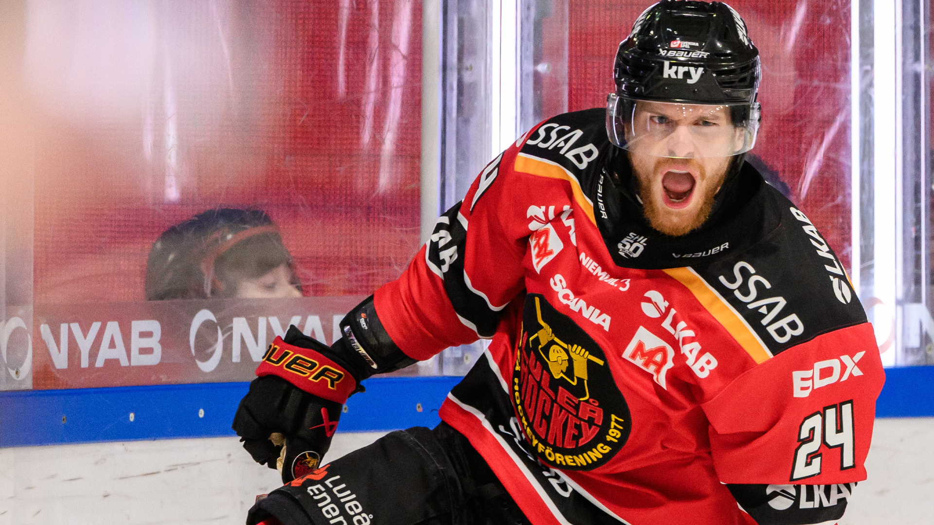 Hockeyspelare i röd och svart tröja sitter på isen under en match med entusiastisk uppsyn. Hockeyspelar i röd och svart tröja poserar framför Linköpings HC:s logga.