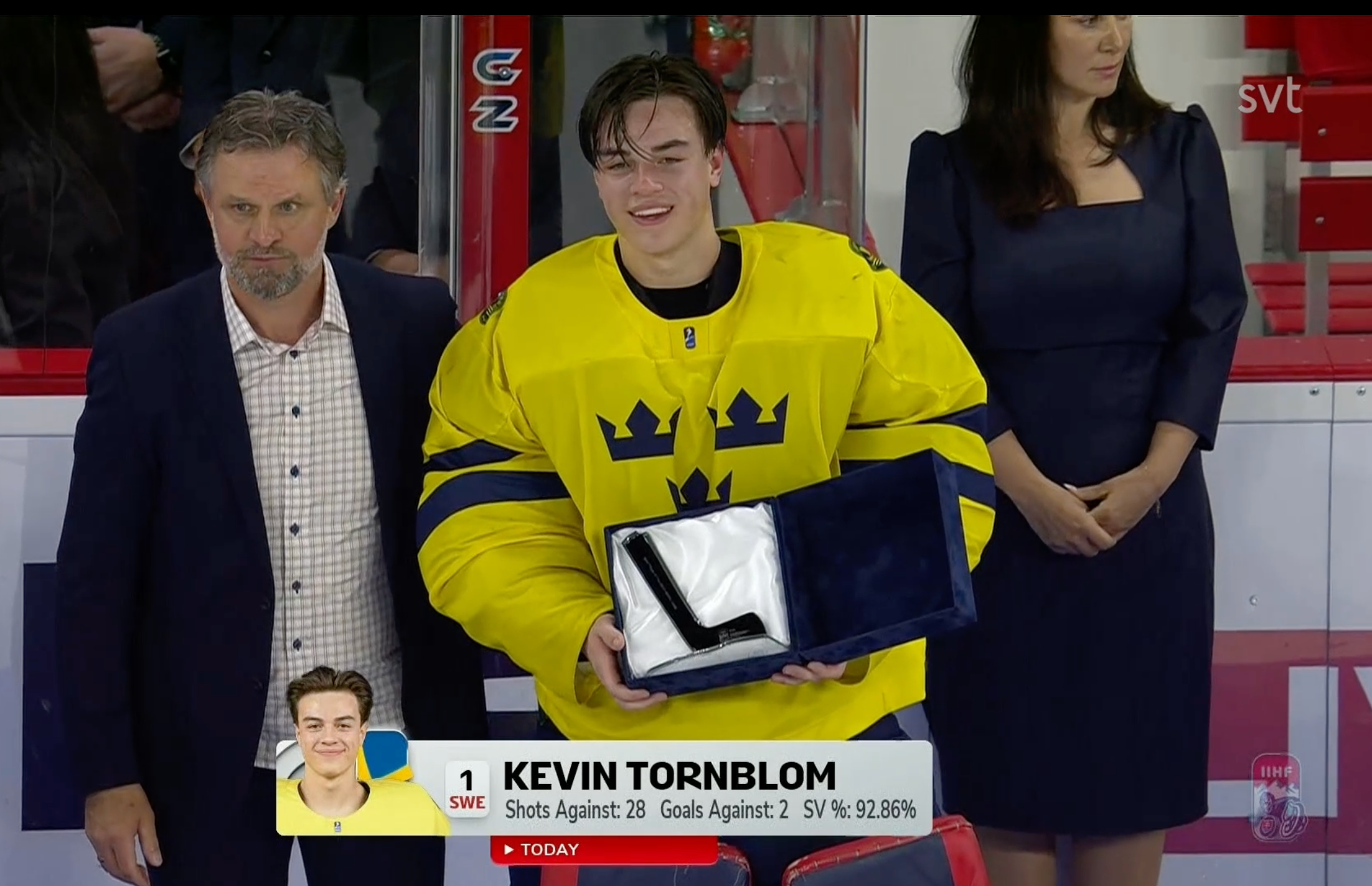 En hockeyspelare i gul svensk landslagströja mottar ett pris eller utmärkelse under en ceremoni. Spelaren omges av officiella personer i formell klädsel.