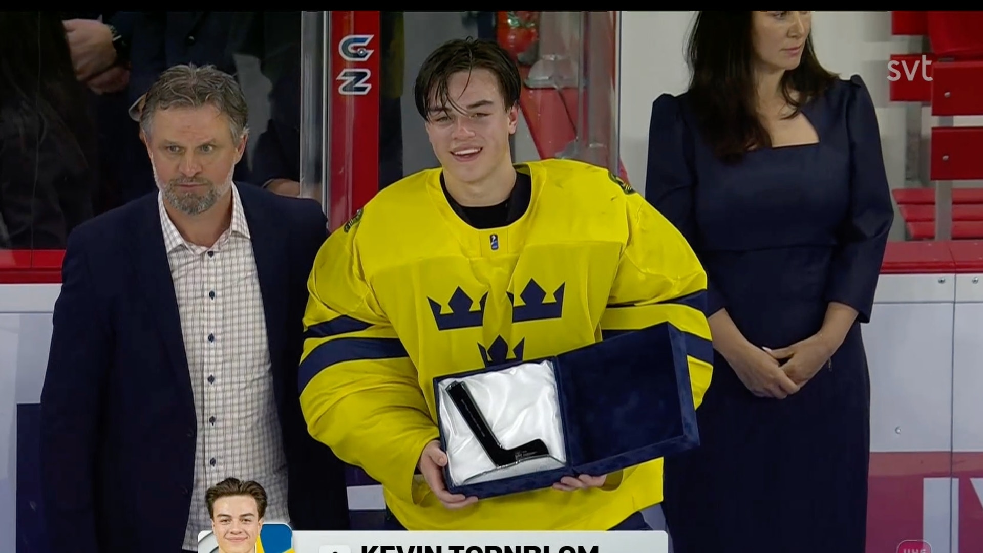En hockeyspelare i gul svensk landslagströja mottar ett pris eller utmärkelse under en ceremoni. Spelaren omges av officiella personer i formell klädsel.