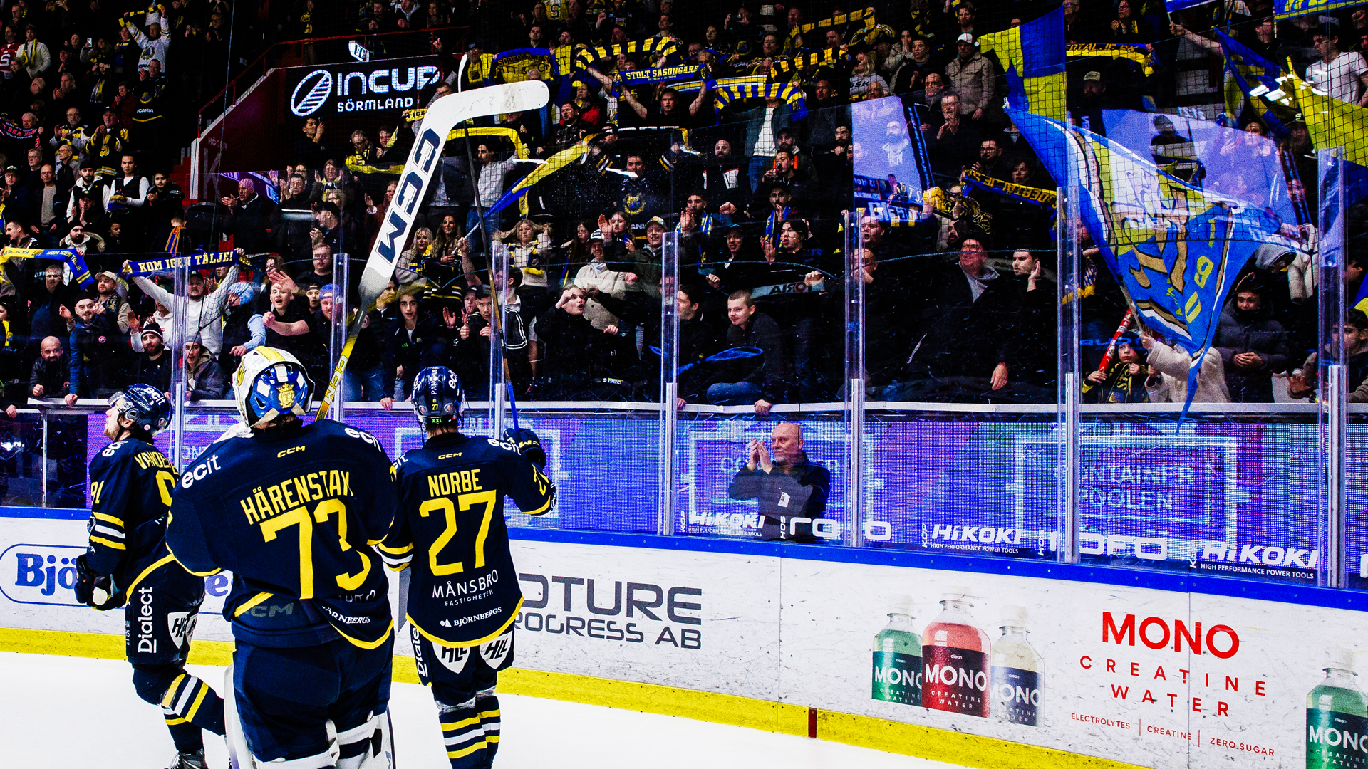 Hockeymatch med spelare i blå och gula tröjor vid bandets kanter. Publik syns i bakgrunden och sponsorlogotyper är synliga längs banden.