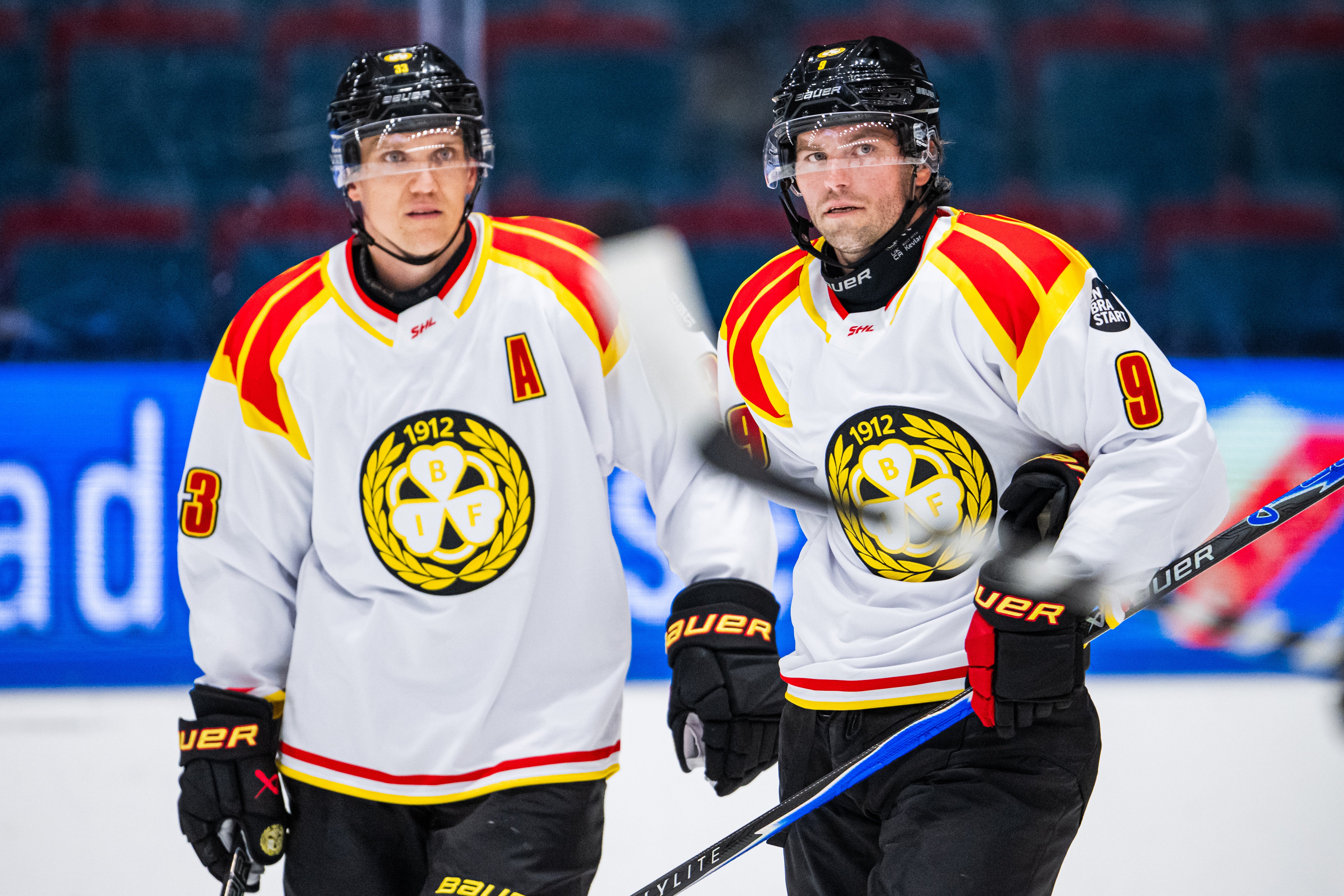 Två ishockeyspelare i vita matchtröjor står på isen under uppvärmning inför en match.
