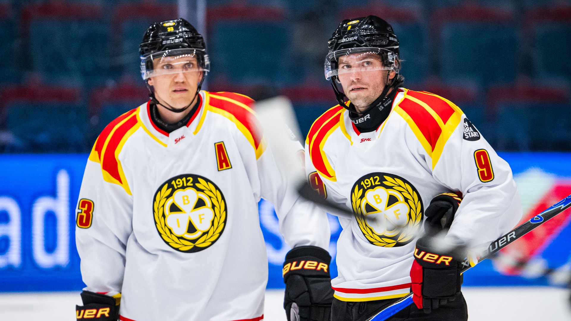 Två ishockeyspelare i vita matchtröjor står på isen under uppvärmning inför en match.