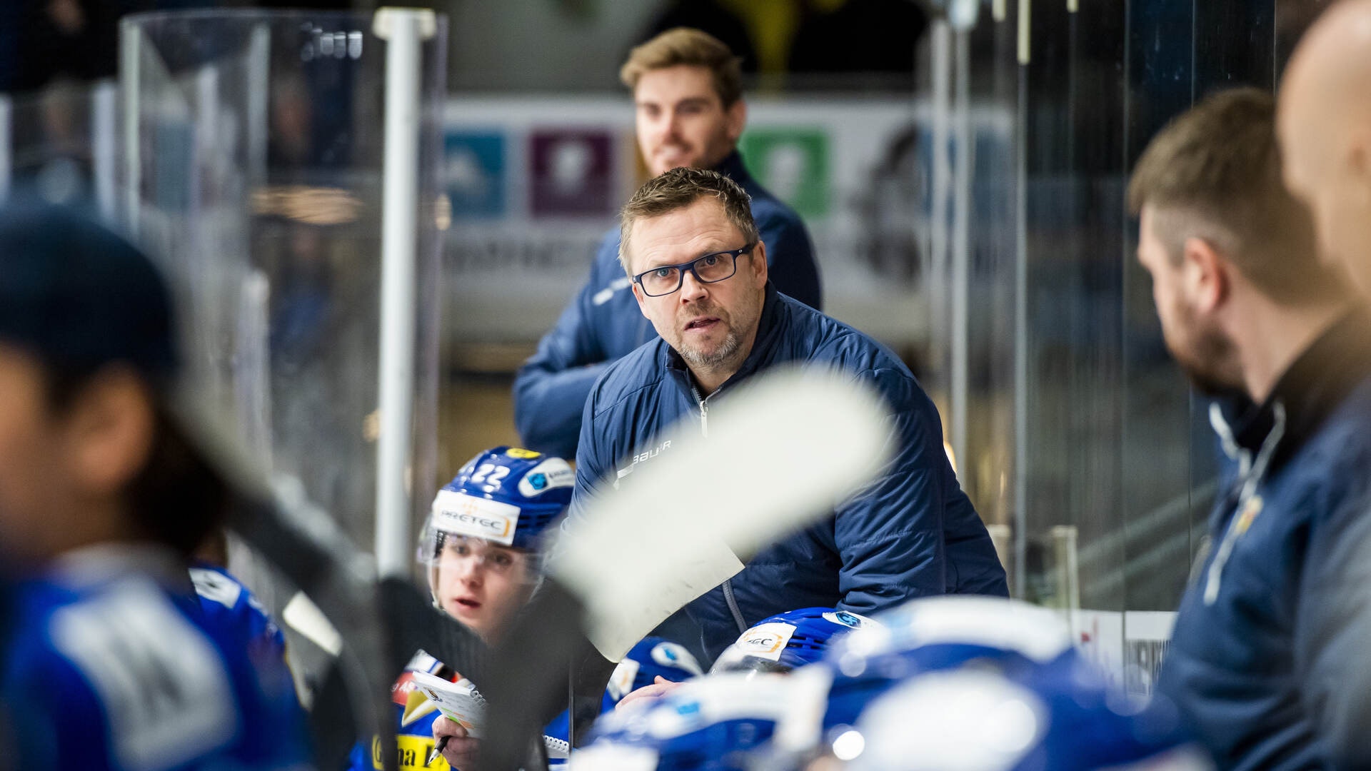 En tränare i blå jacka sitter vid ishockeybänken och observerar spelet. Han är omgiven av spelare i blå och vita uniformer.