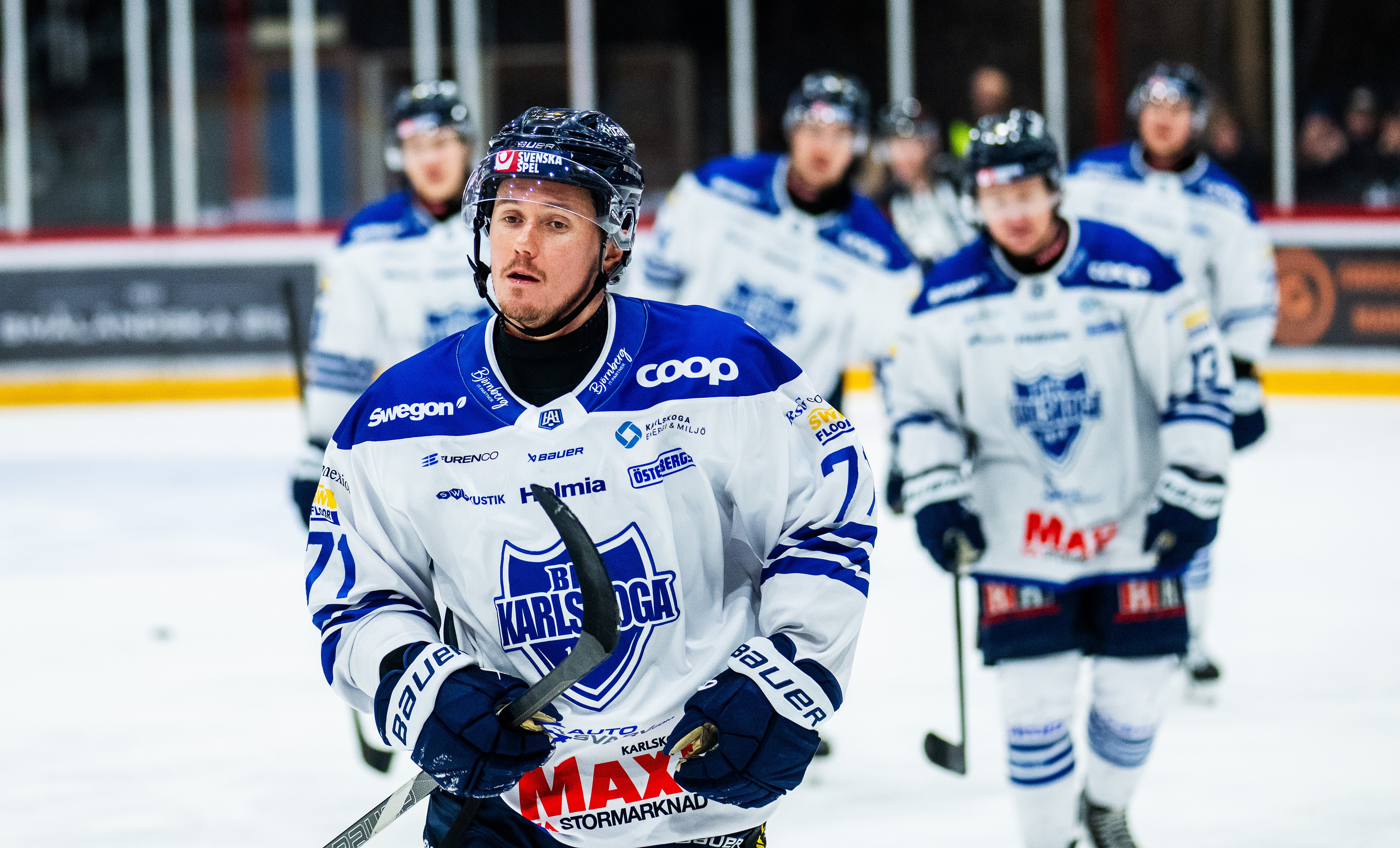 Ishockeyspelare i blå och vit tröja spelar på isen under en match, med lagkamrater i bakgrunden.