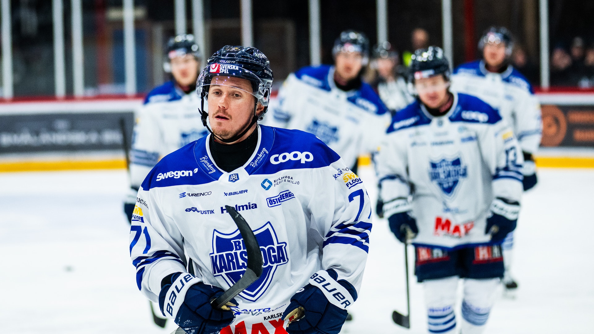 Ishockeyspelare i blå och vit tröja spelar på isen under en match, med lagkamrater i bakgrunden.