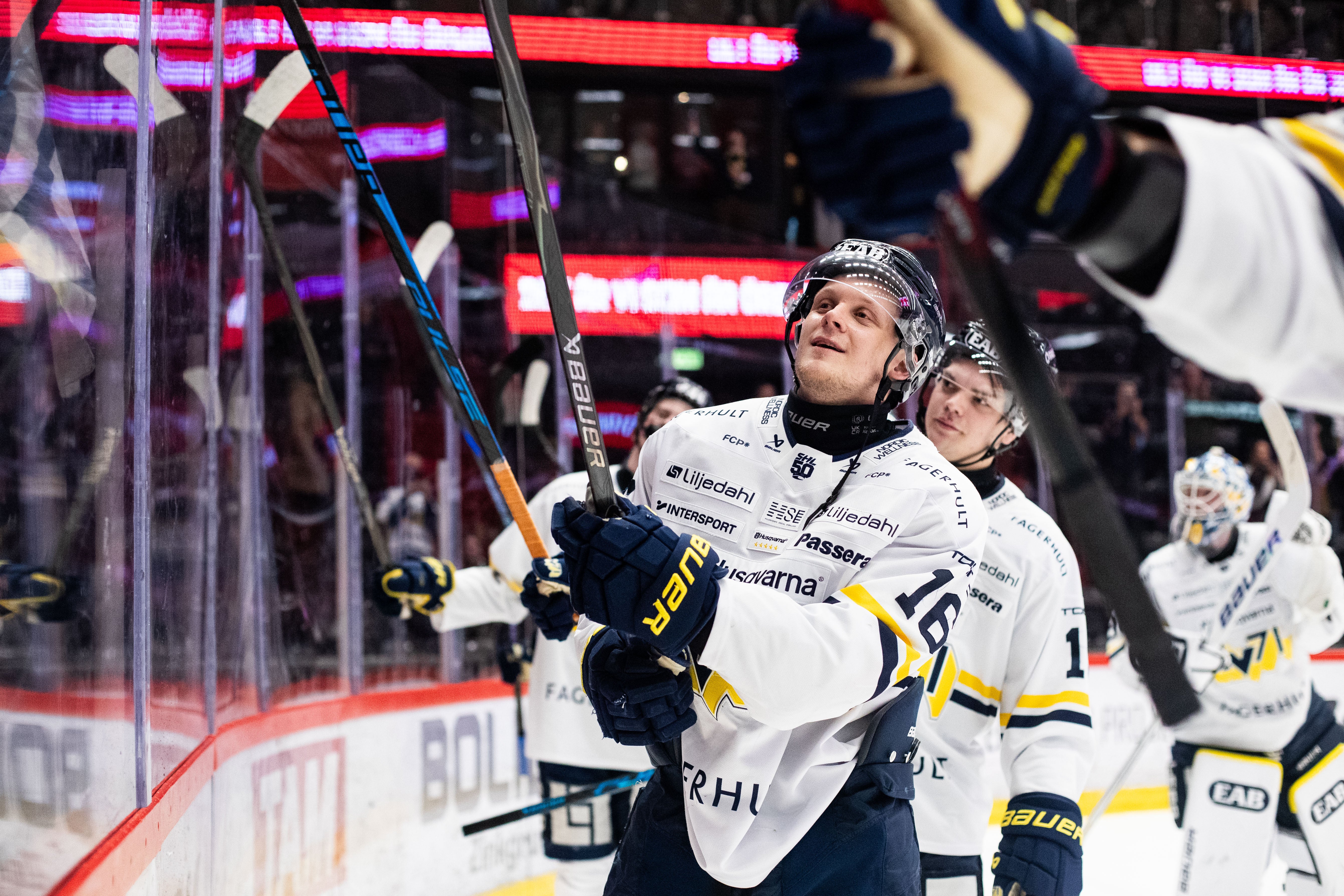 Två ishockeyspelare i vita och gula tröjor firar under en match. De håller upp sina klubbor mot publiken och ser glada ut. I bakgrunden syns röda neonljus från arenan.