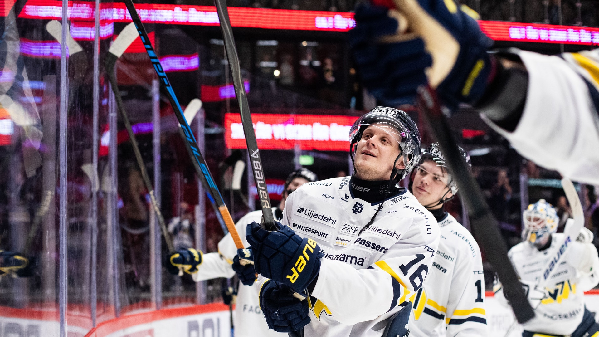 Två ishockeyspelare i vita och gula tröjor firar under en match. De håller upp sina klubbor mot publiken och ser glada ut. I bakgrunden syns röda neonljus från arenan.